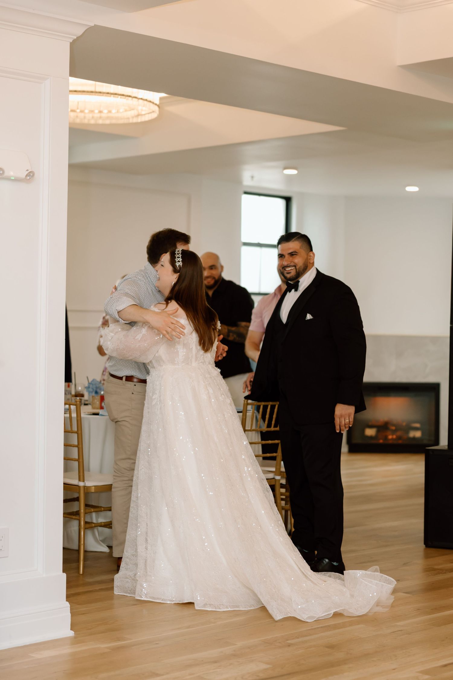 Bride embraces guest while groom smiles during wedding reception entrance at elegant venue.