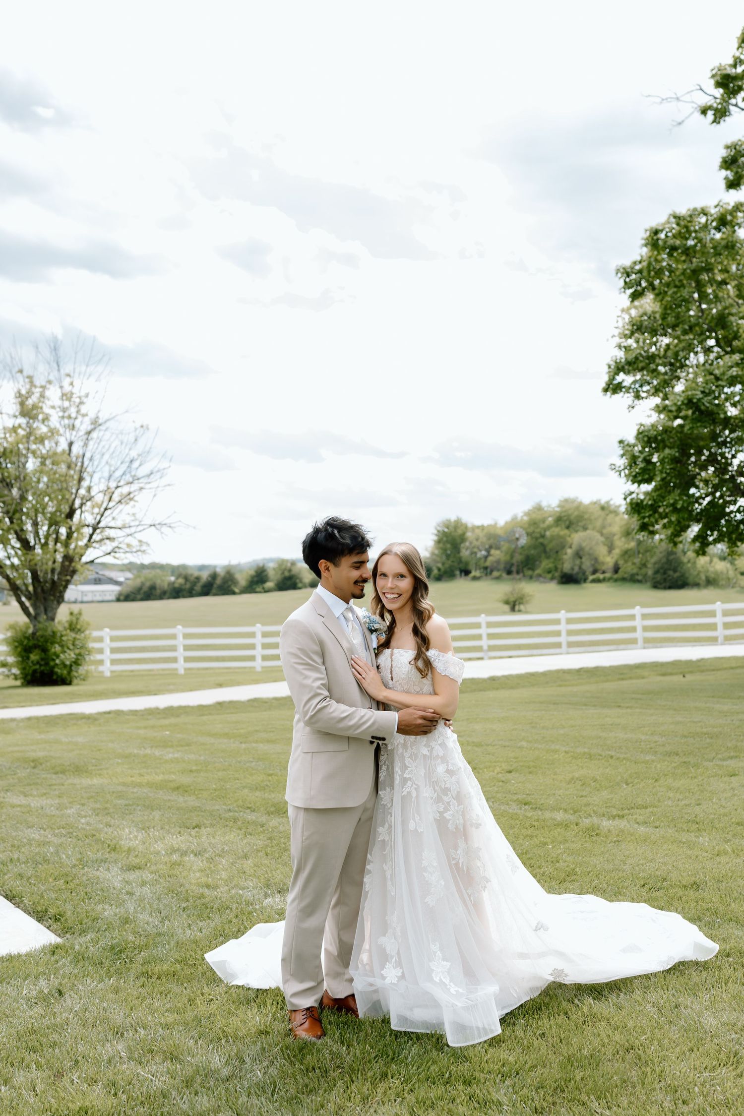 A bride and groom embrace on a lush green lawn, she in a floral gown with train, he in a beige suit, with white fences behind.