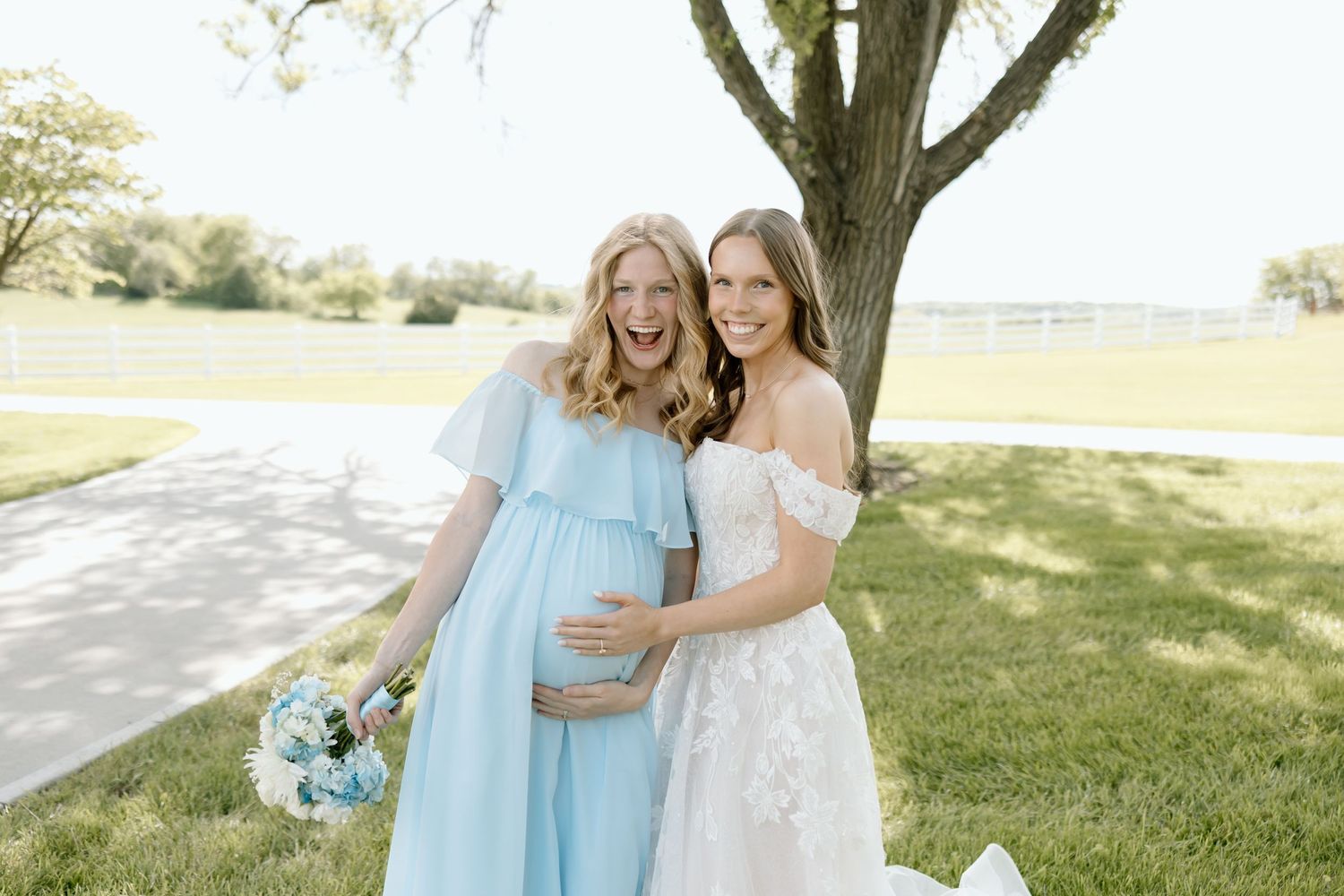 Pregnant woman in blue dress poses with bride in white lace gown outdoors near a tree on a sunny day.
