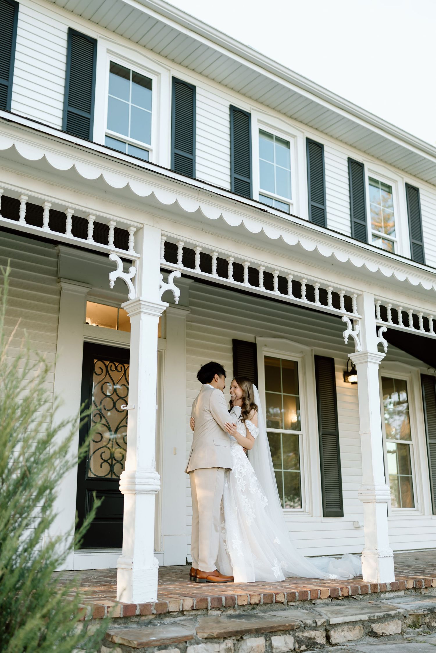 A couple in wedding attire shares a romantic moment on the porch of a charming white colonial home.
