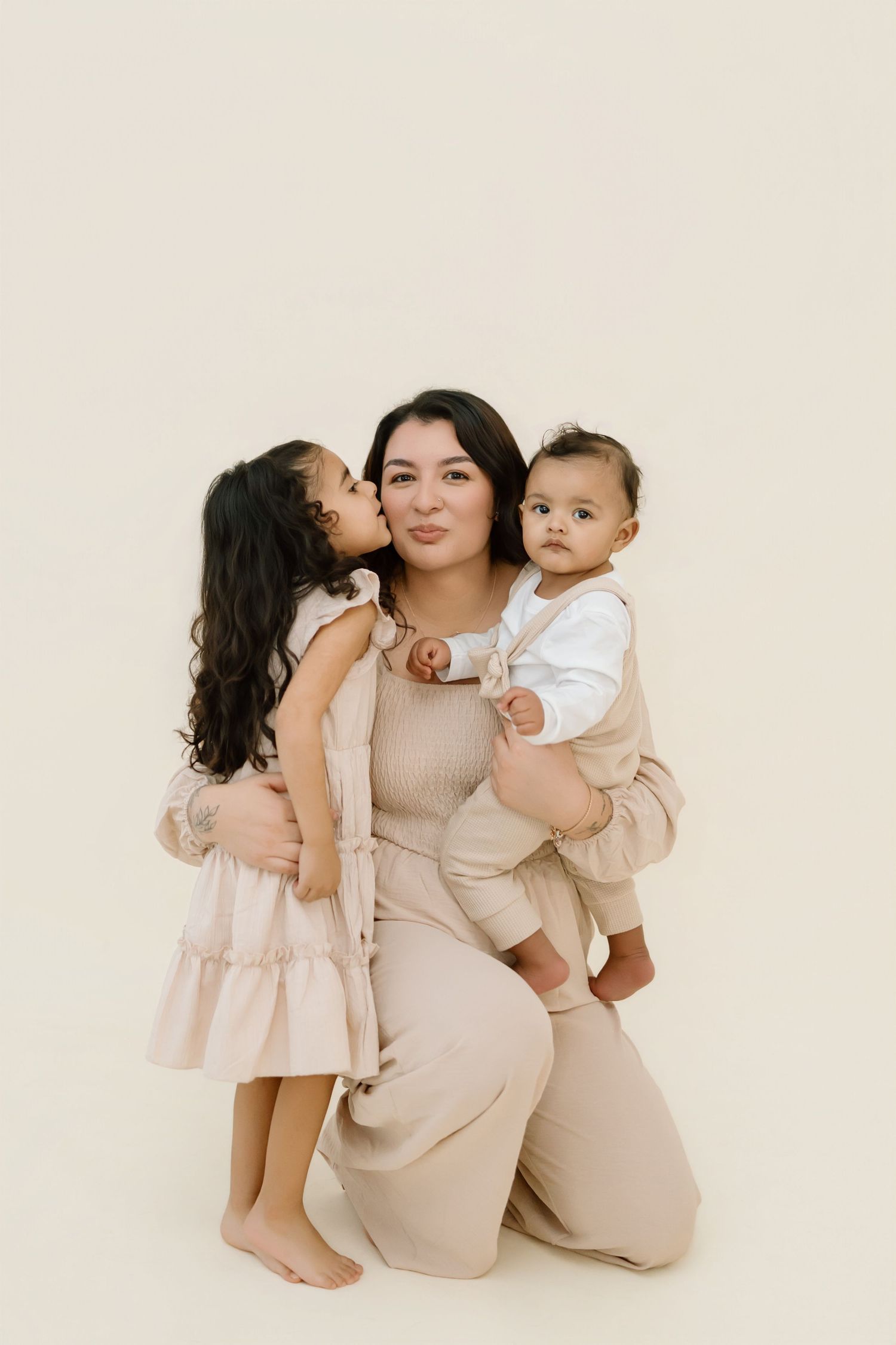 A family portrait in neutral tones featuring a mother with two young children against a light beige background.