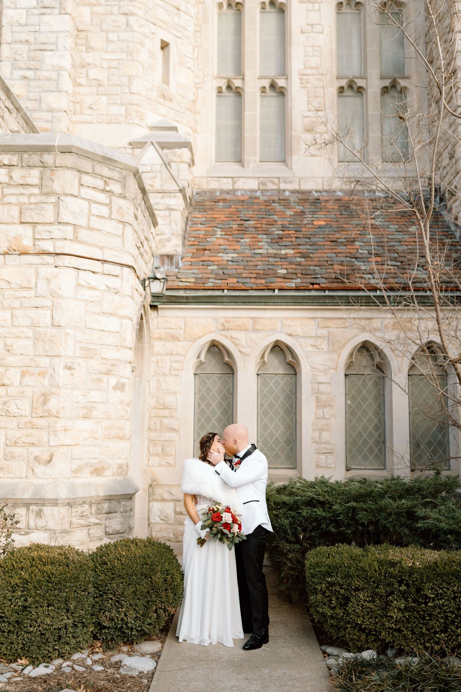 A romantic winter wedding moment captured outside a historic stone church with Gothic architecture and bare trees.