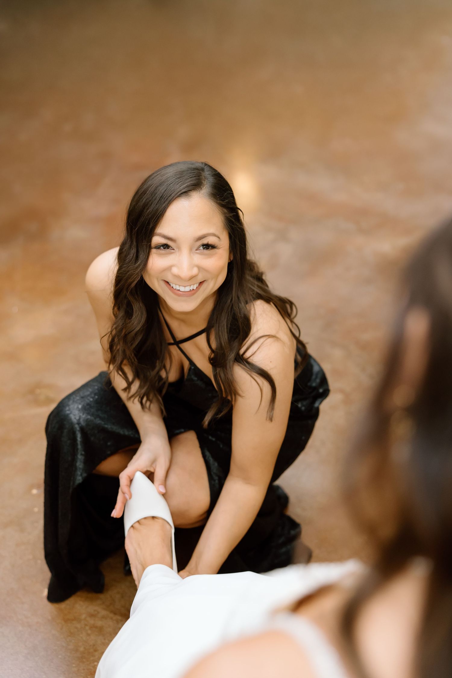 A woman in a black dress smiles warmly while sitting on the floor during a portrait session.