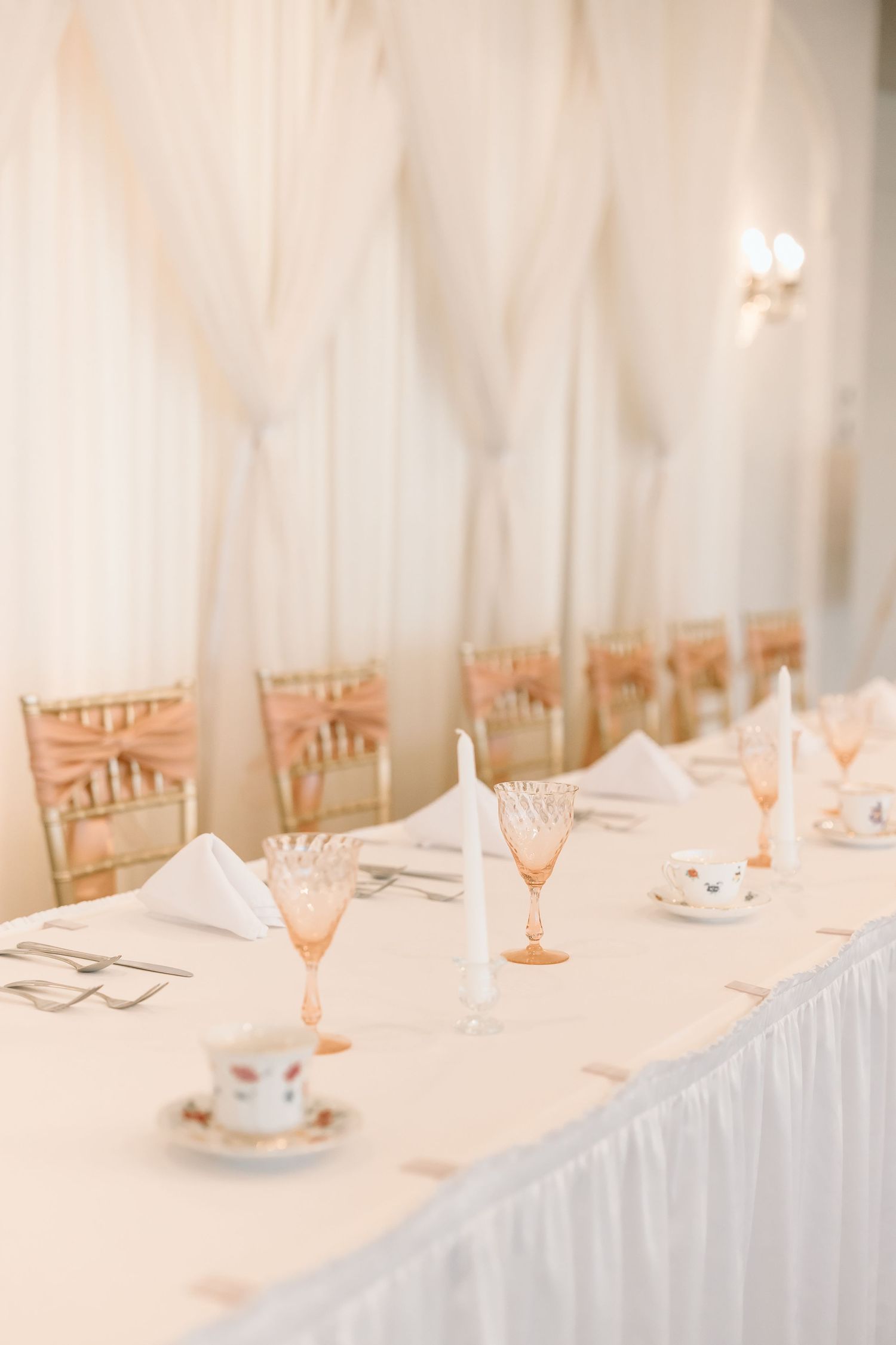 Elegant wedding reception table setting with pink champagne glasses and white linens against a light draped backdrop.