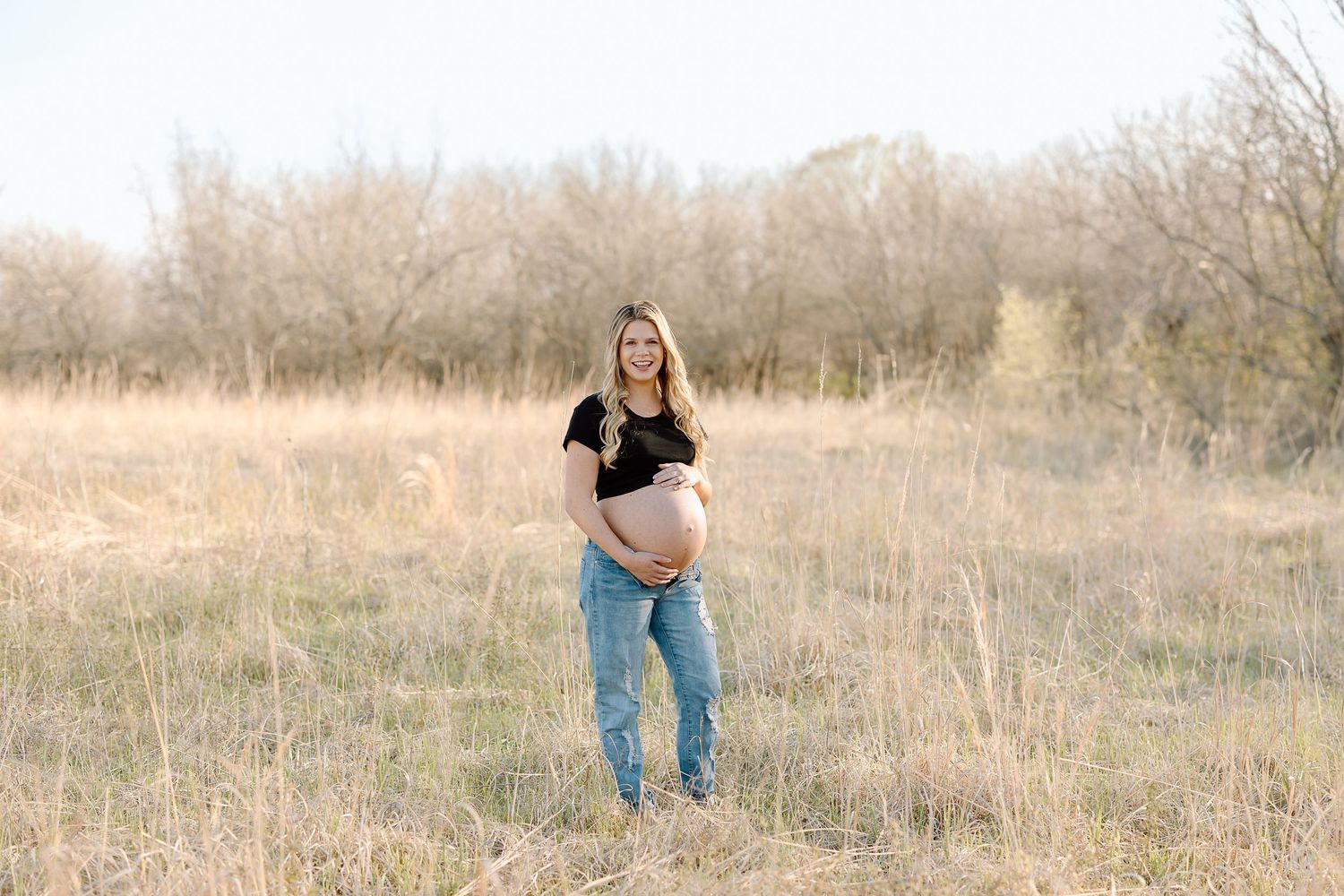 Expectant mother in jeans and black top stands in golden field during sunset maternity photoshoot.