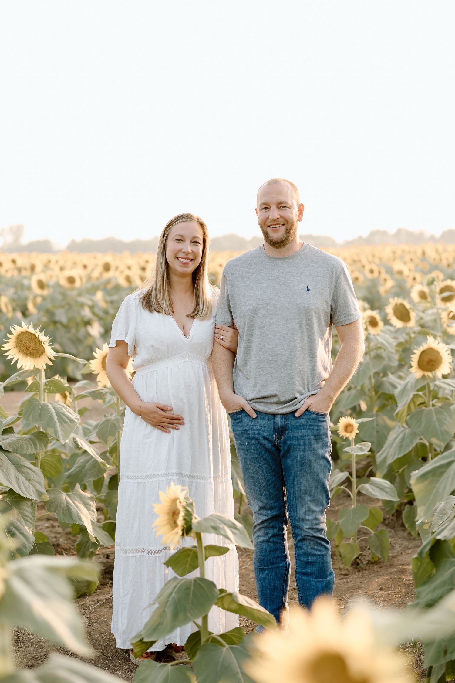 A couple poses together in a sunlit sunflower field during golden hour, wearing casual white and grey attire.