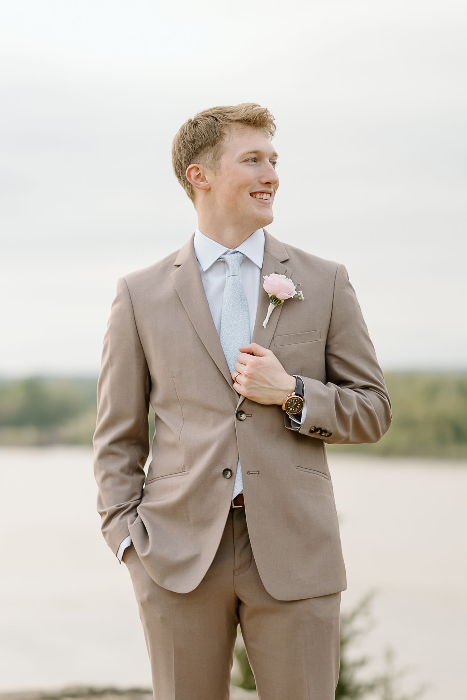 A groomsman in a tan suit and light blue tie stands on a beach smiling with a pink boutonniere.