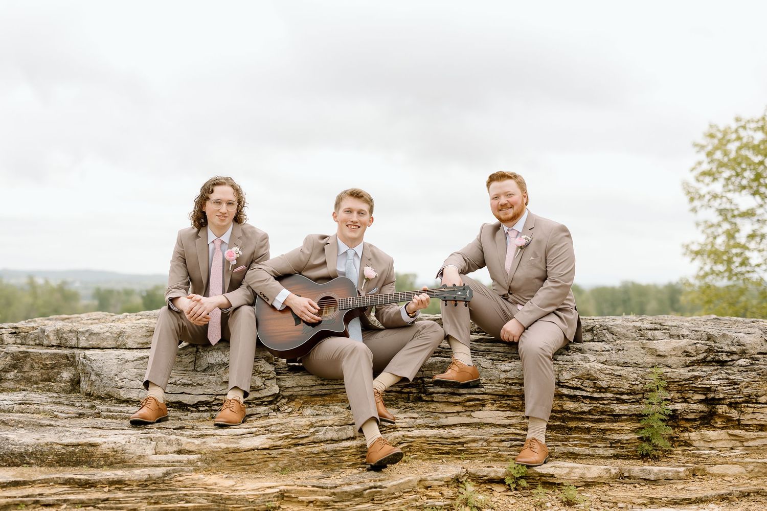 A group wearing gray suits and brown shoes sits casually on a rustic log wall in an outdoor setting.