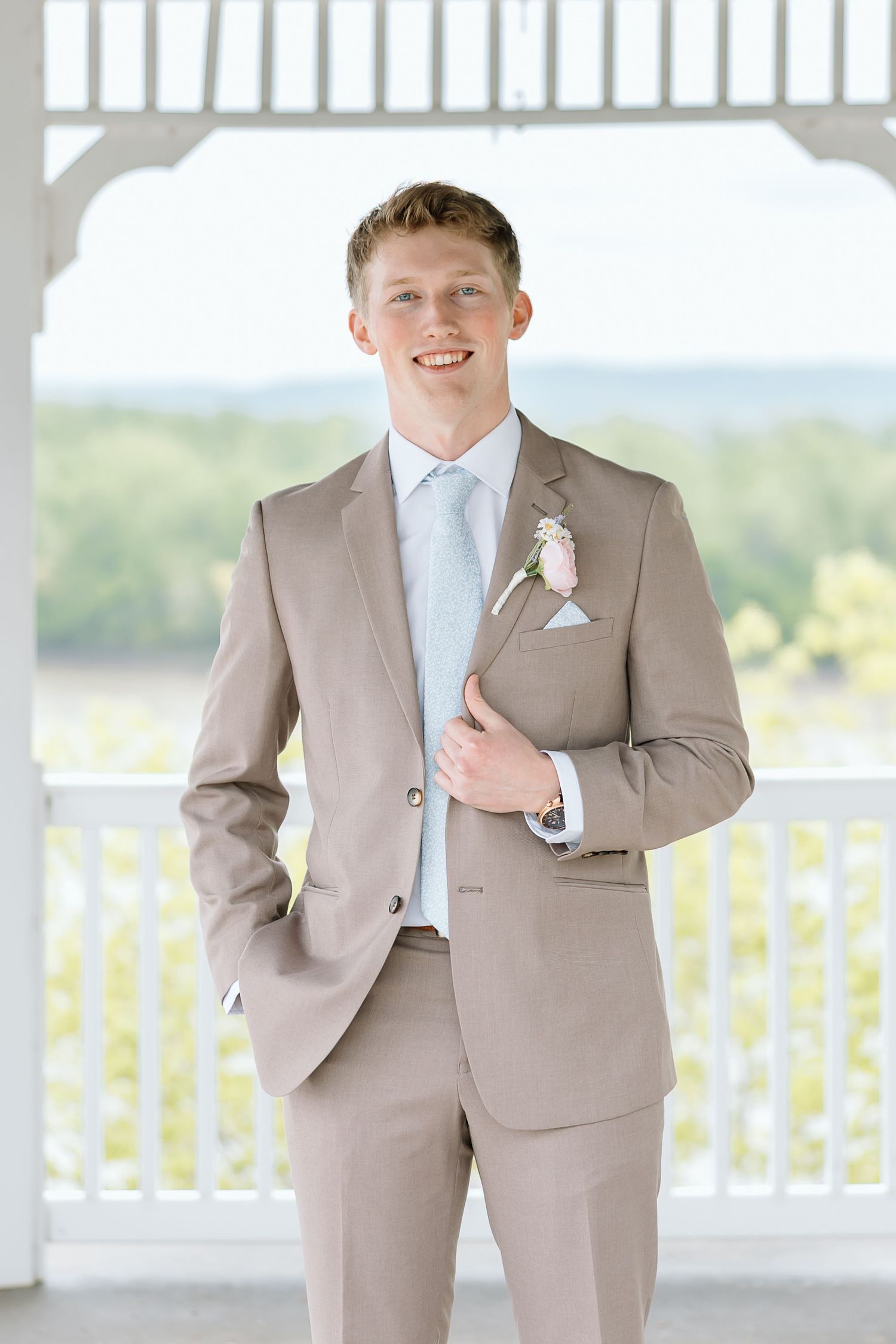 A groom in a light tan suit poses for formal wedding portraits on a white porch with railing.