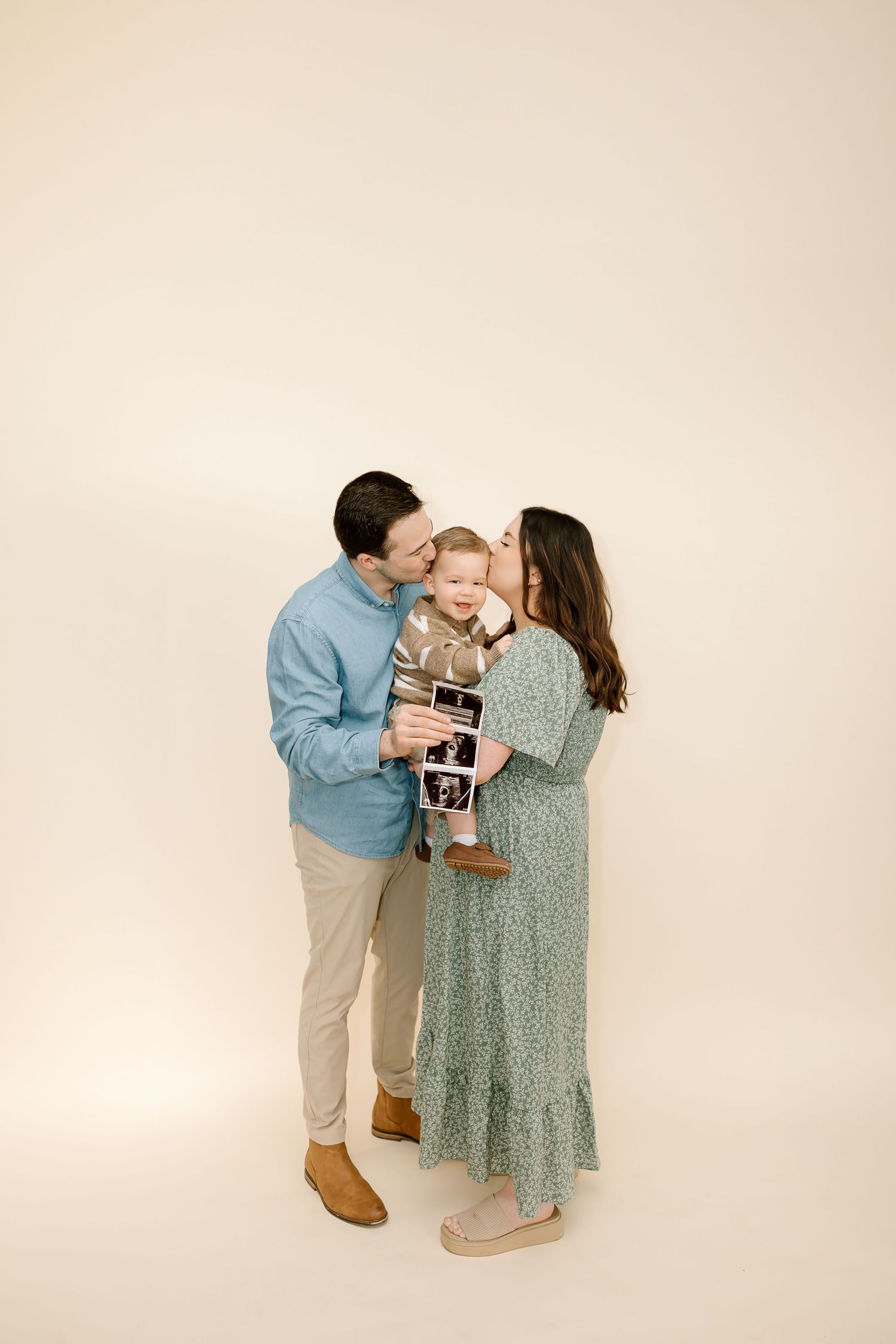 A couple in matching blue and green outfits share a tender moment while holding ultrasound photos in a studio setting.