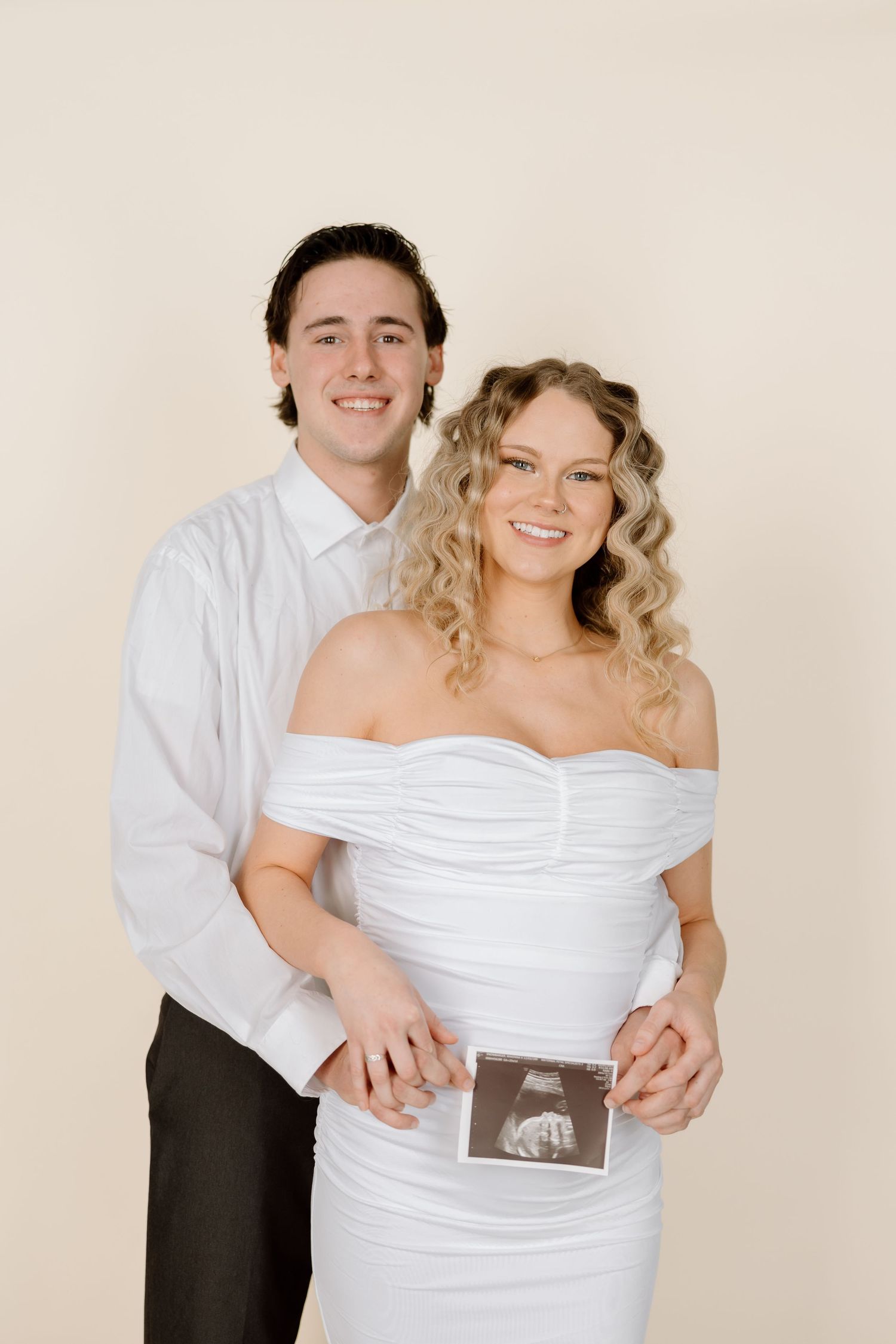Couple in white and black attire share maternity photos while holding ultrasound image against beige backdrop.