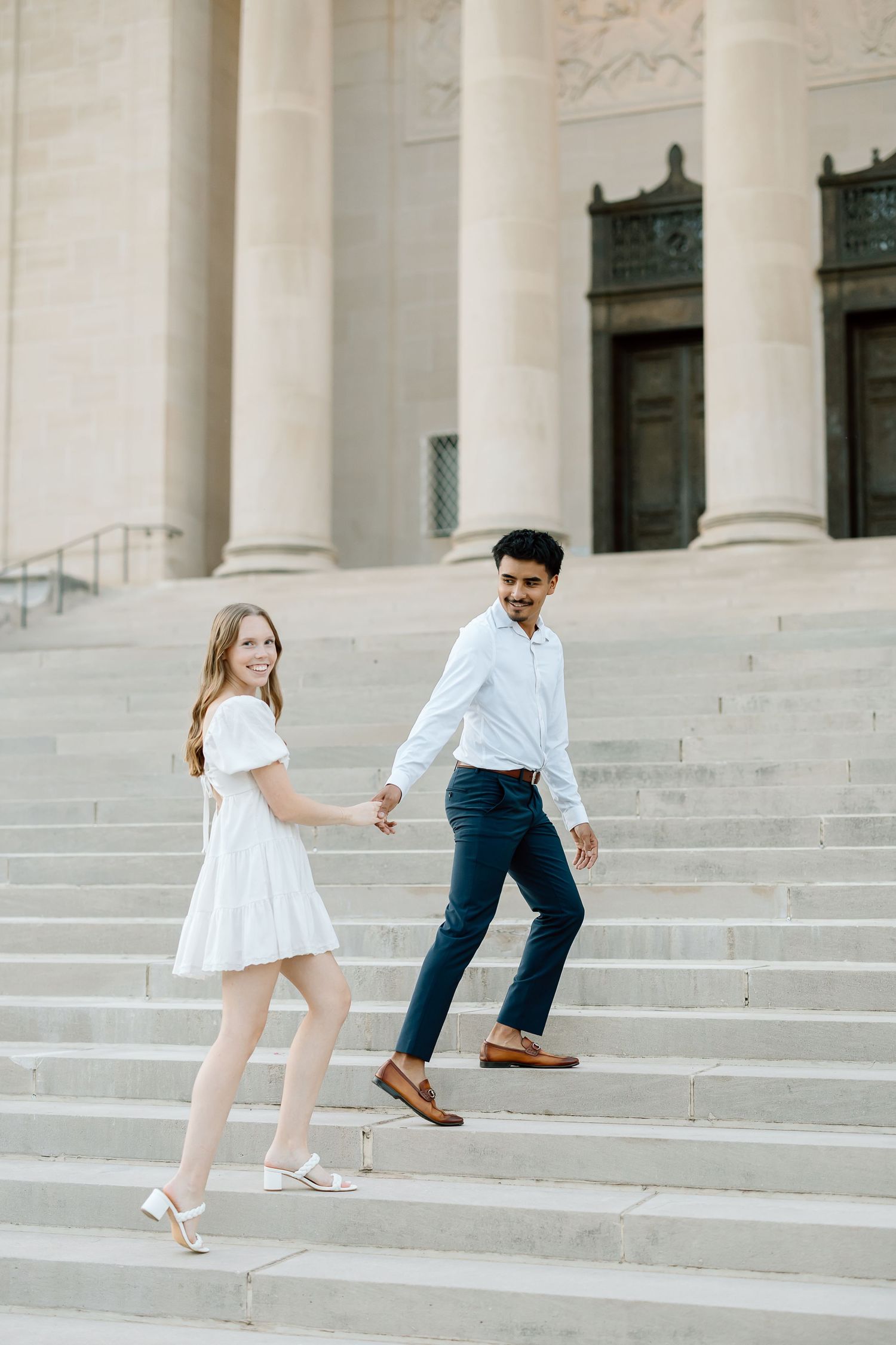 A couple in white and blue outfits playfully walks up stone steps in a romantic engagement photo sequence.