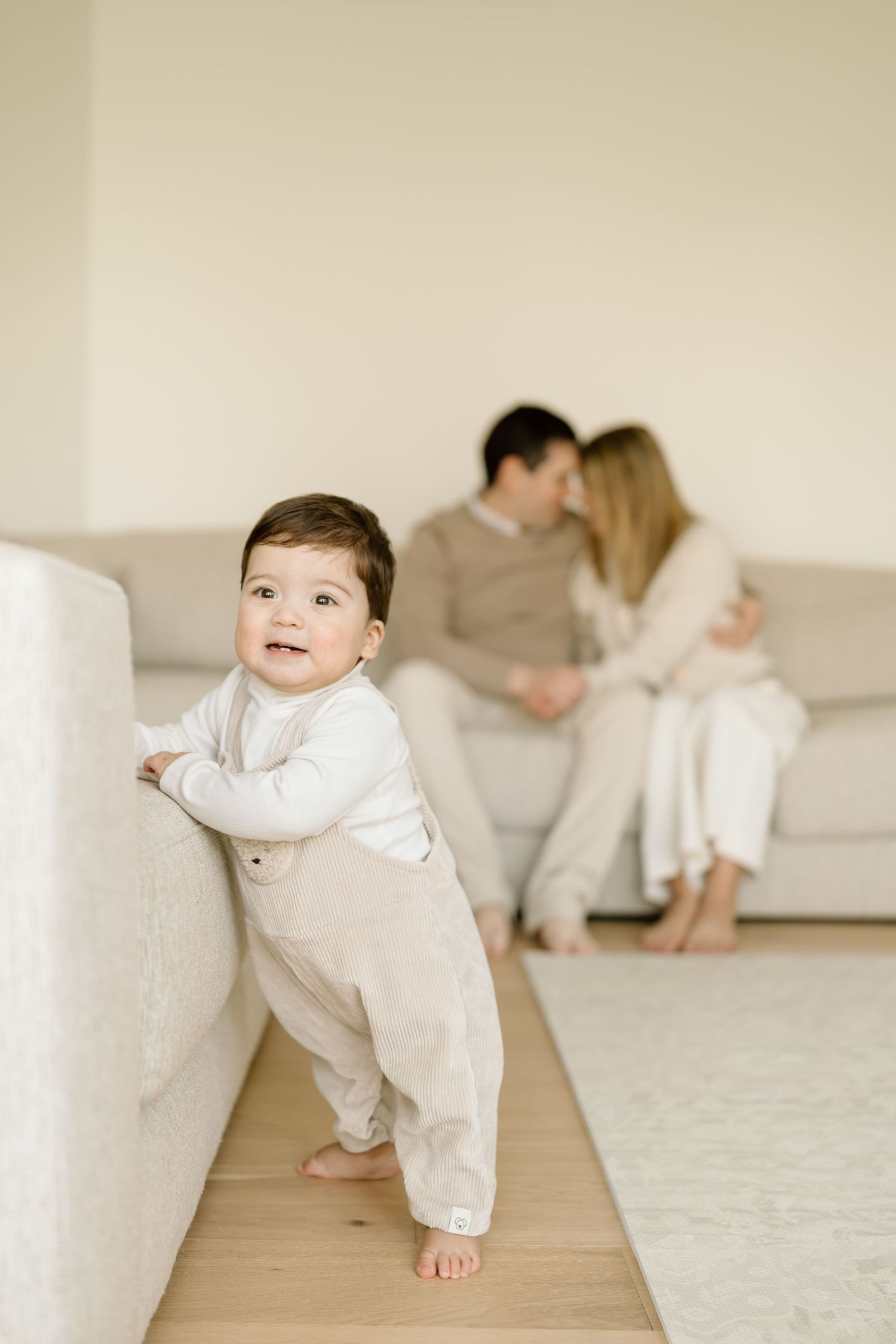 A baby in white clothing crawls across a light wooden floor with adults sitting on a couch in the background.