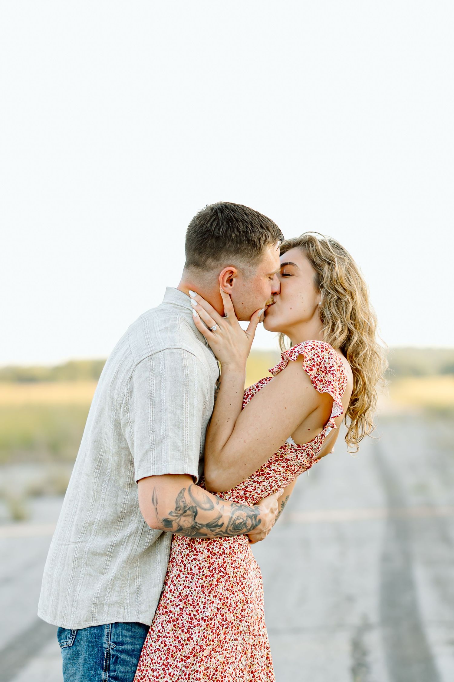 Romantic couple sharing tender kisses at sunset on a sandy beach wearing white shirt and floral dress.