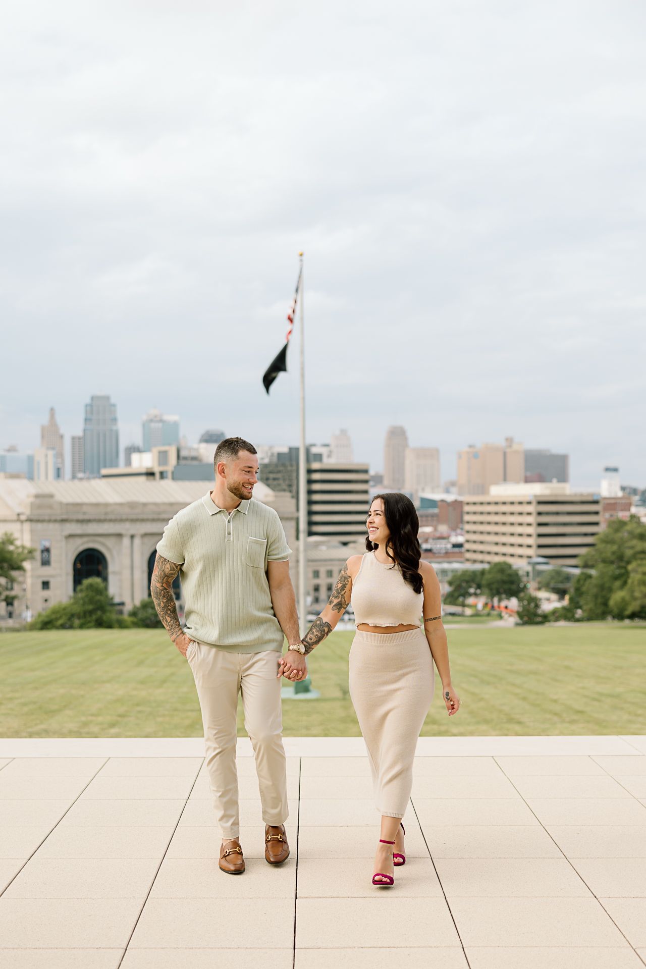 A couple walks together on a paved plaza with the Kansas City skyline visible in the background.