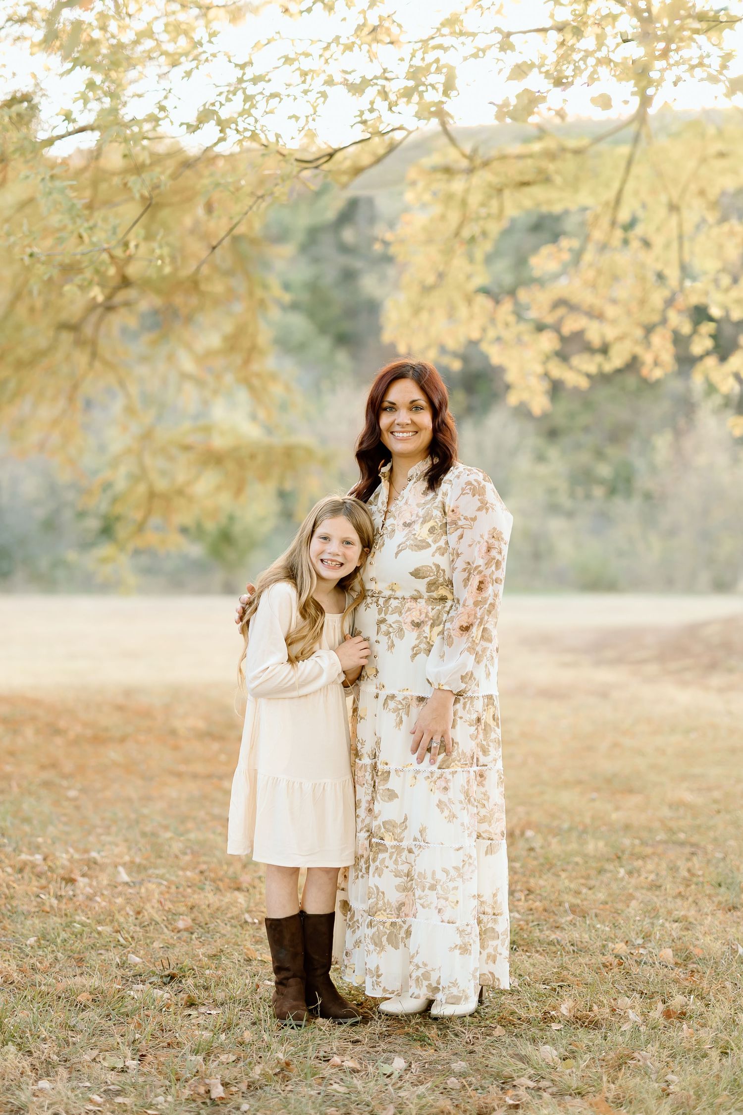Two people in light colored dresses stand together on autumn grass with golden trees in the background.