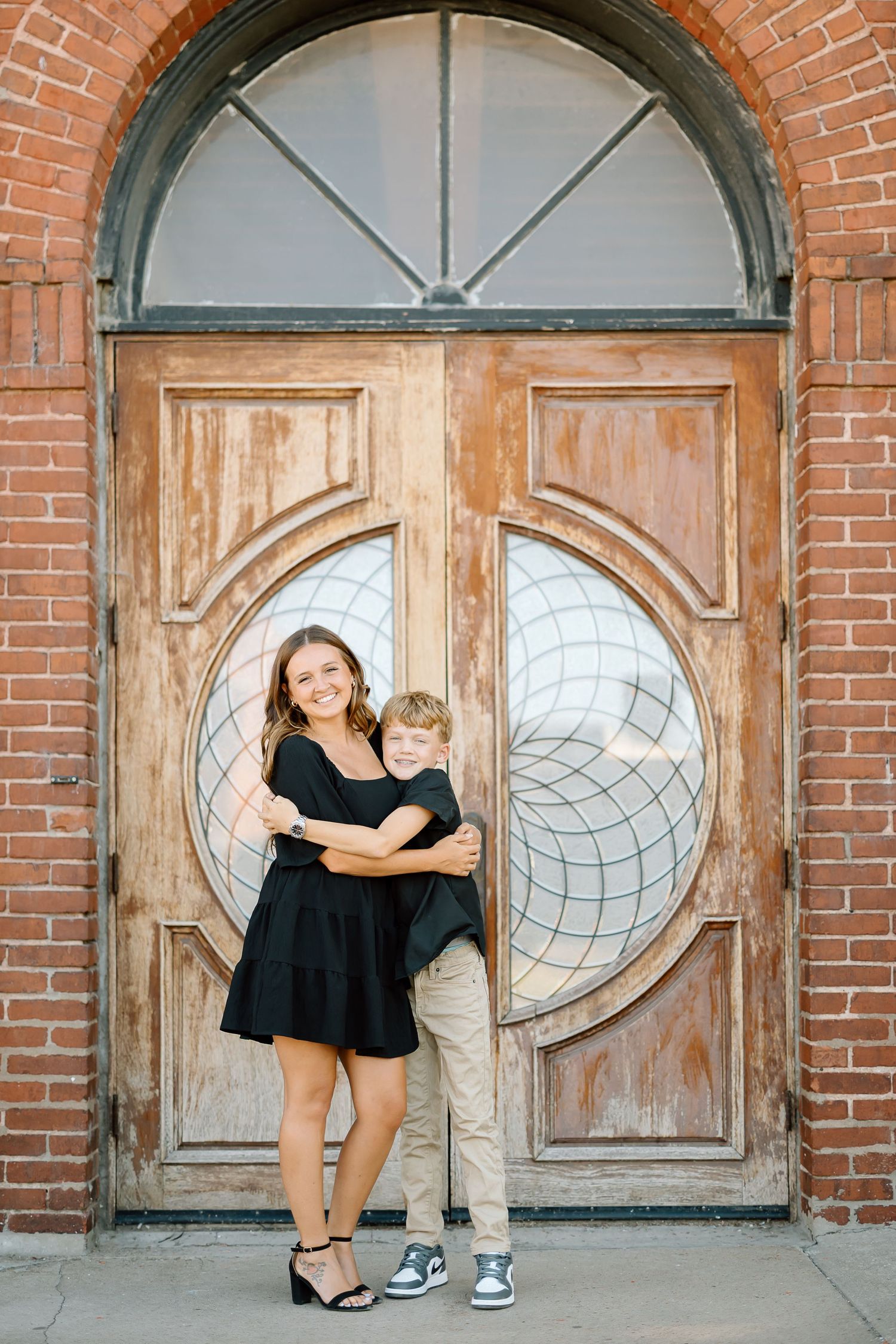 A couple in formal and casual attire poses in front of vintage wooden doors with circular glass windows and brick walls.