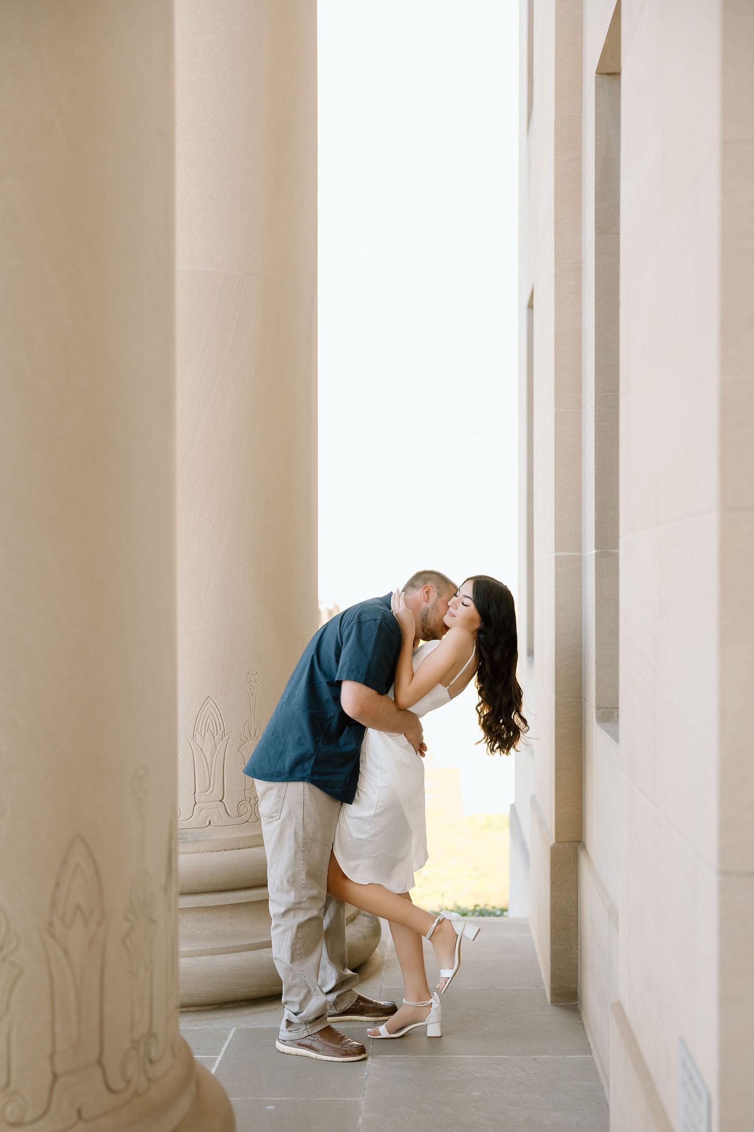 A romantic couple shares a tender kiss in an elegant hallway with white columns and natural lighting.