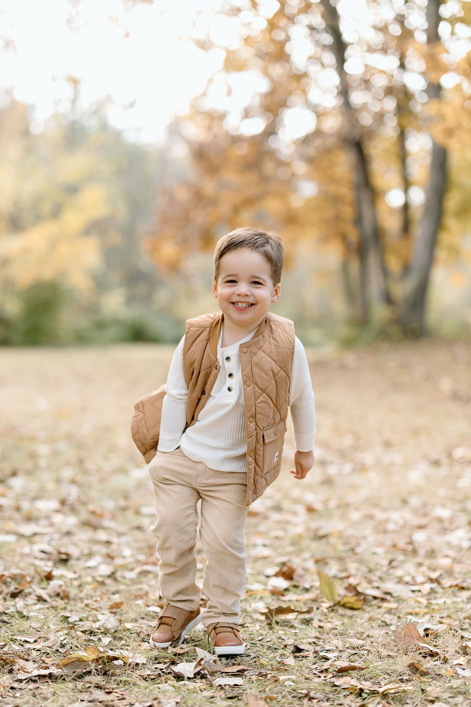 A young child in a brown vest and khaki pants plays in fallen autumn leaves on a sunny day.