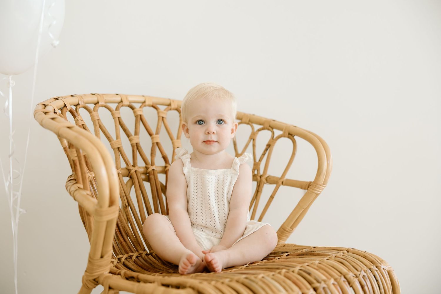 Baby wearing pink bloomers sits in vintage wicker peacock chair against white background.