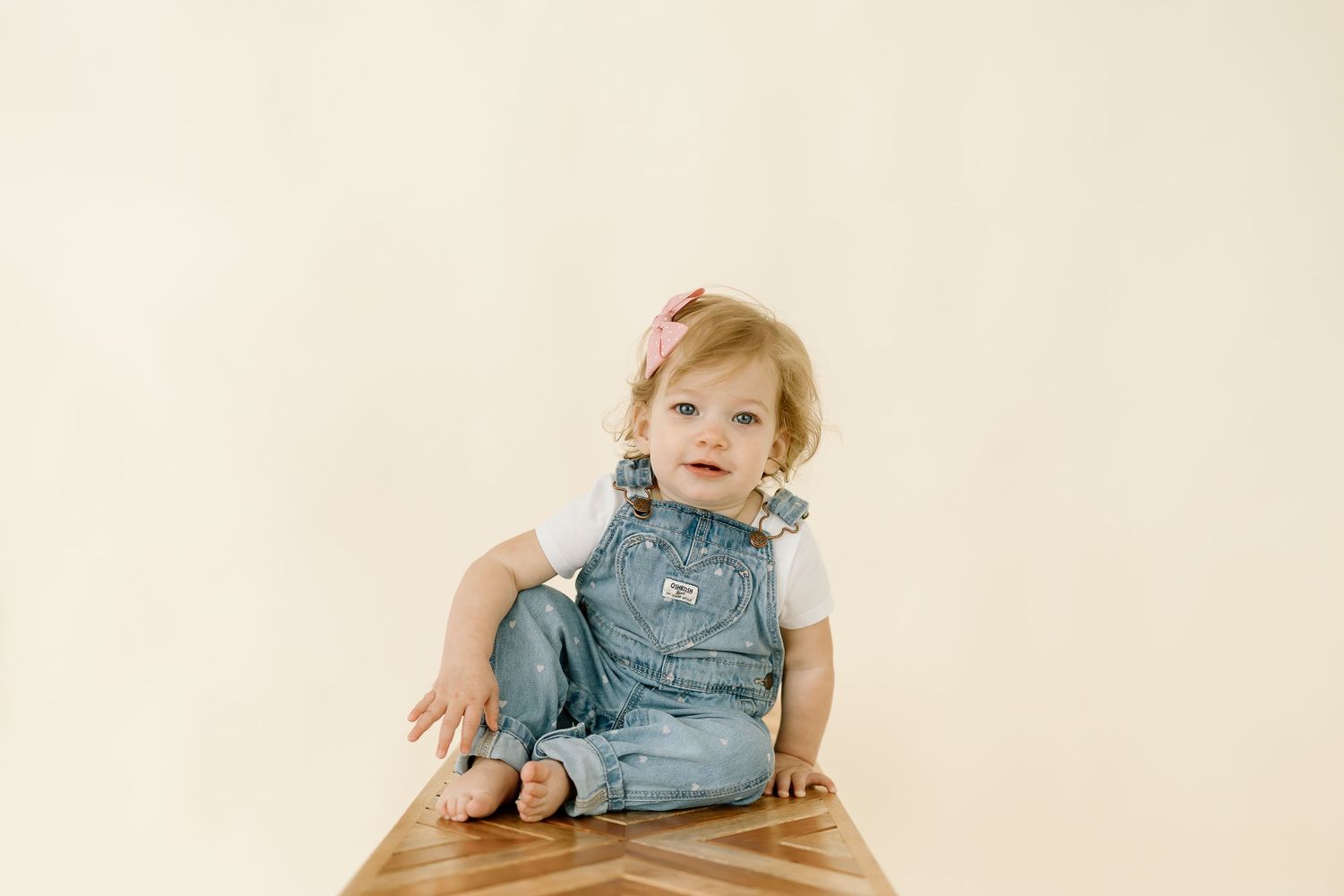 A toddler in denim overalls sits and plays on a light beige background in a series of candid poses.