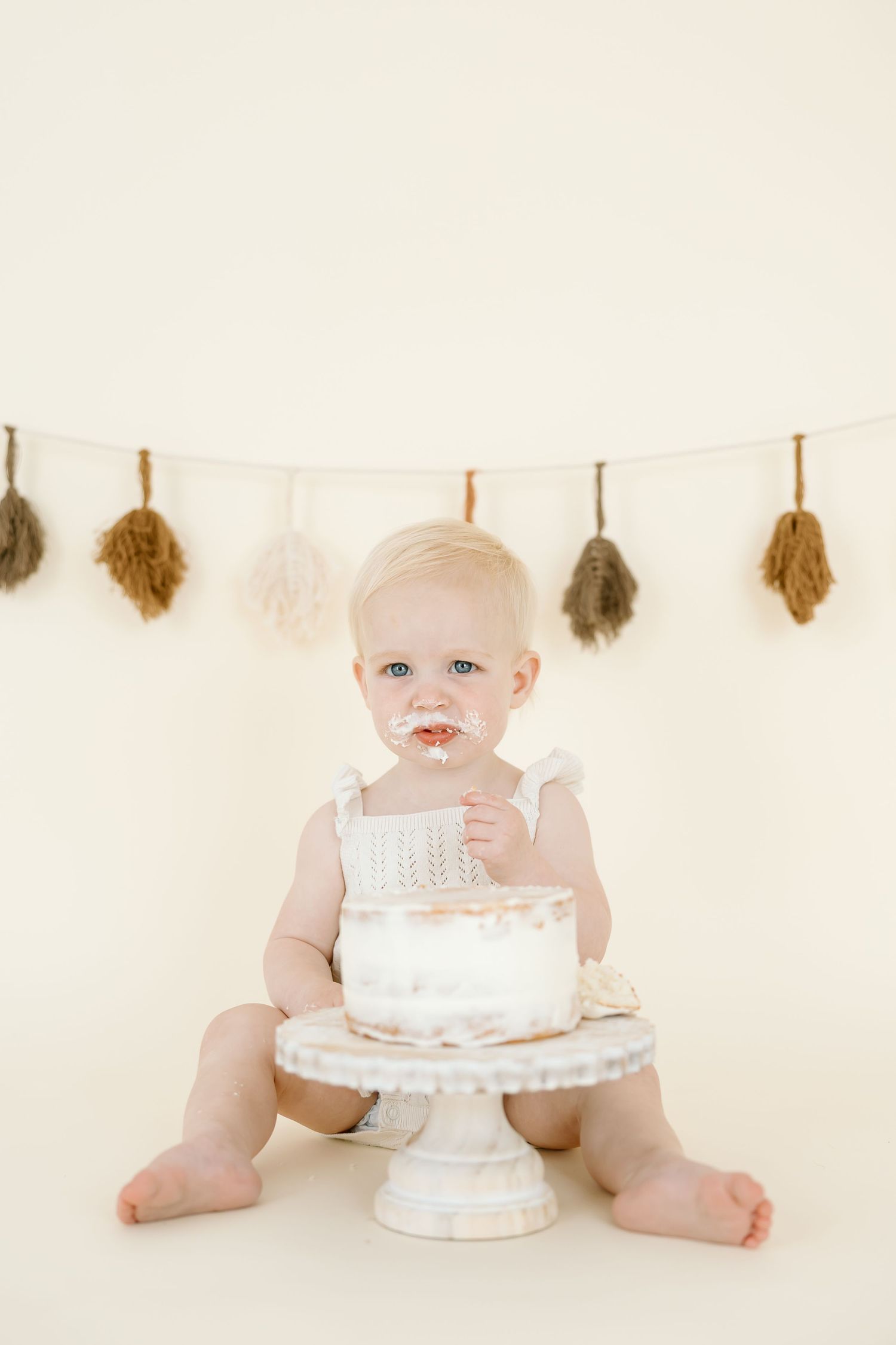 A small child sits with a white cake on a pedestal stand against a neutral backdrop with dried leaves hanging above.