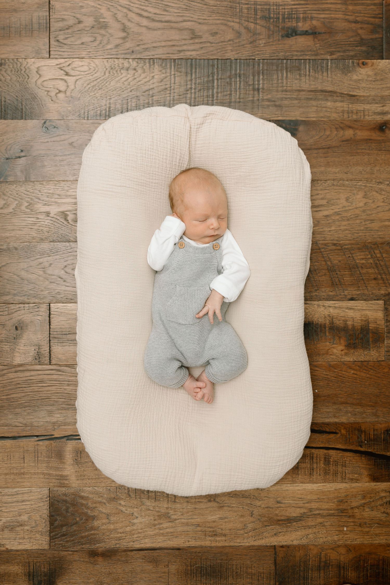 Newborn baby in gray onesie sleeping on cream cushion against rustic wooden background.