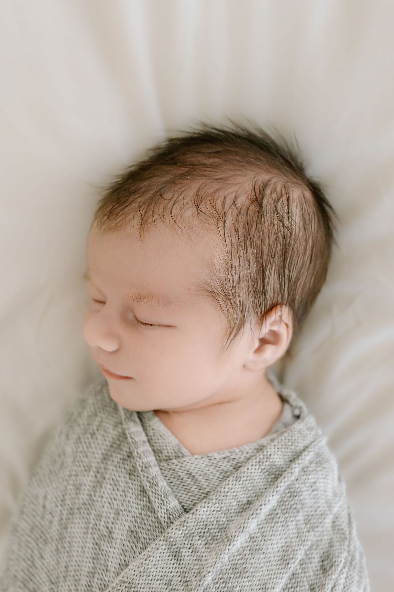 A sleeping newborn baby wrapped in a soft gray blanket rests peacefully on white bedding.