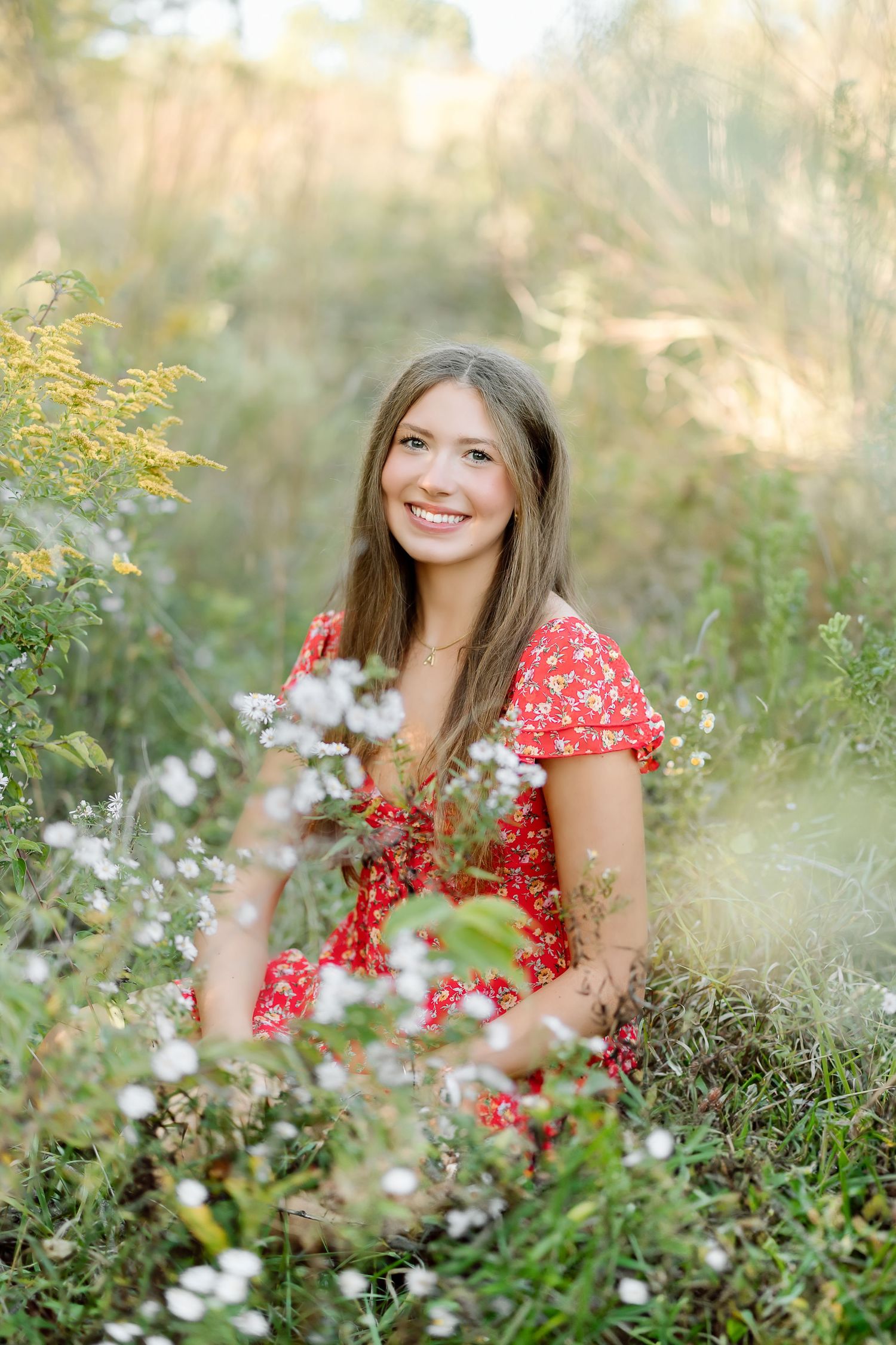 A person in a red floral dress smiles while sitting among wildflowers in a dreamy outdoor setting.