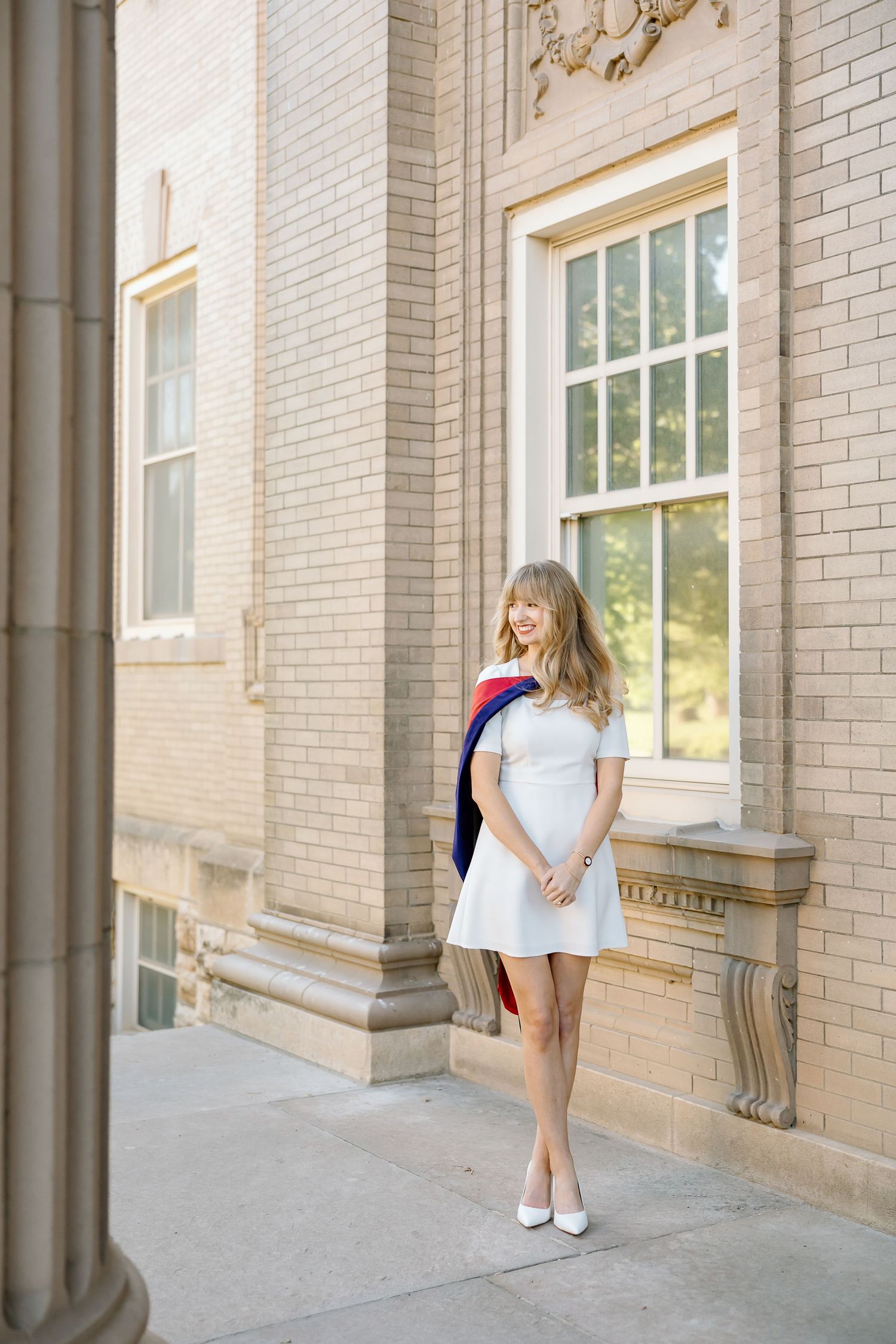 A person in a white dress stands against a light brick wall near large windows in a classic architectural setting.