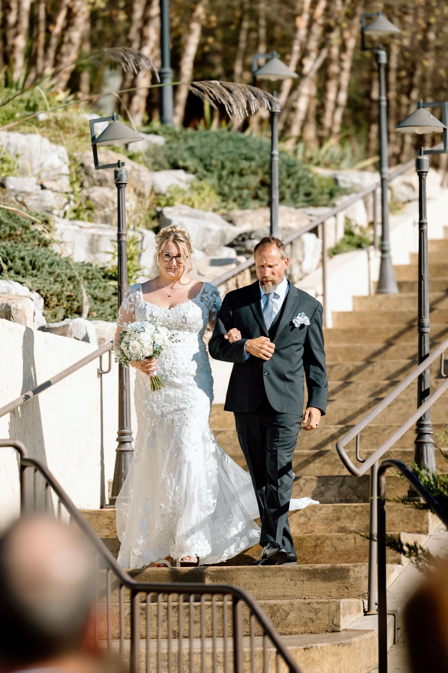 A couple in wedding attire descends an outdoor stone staircase surrounded by trees and greenery.
