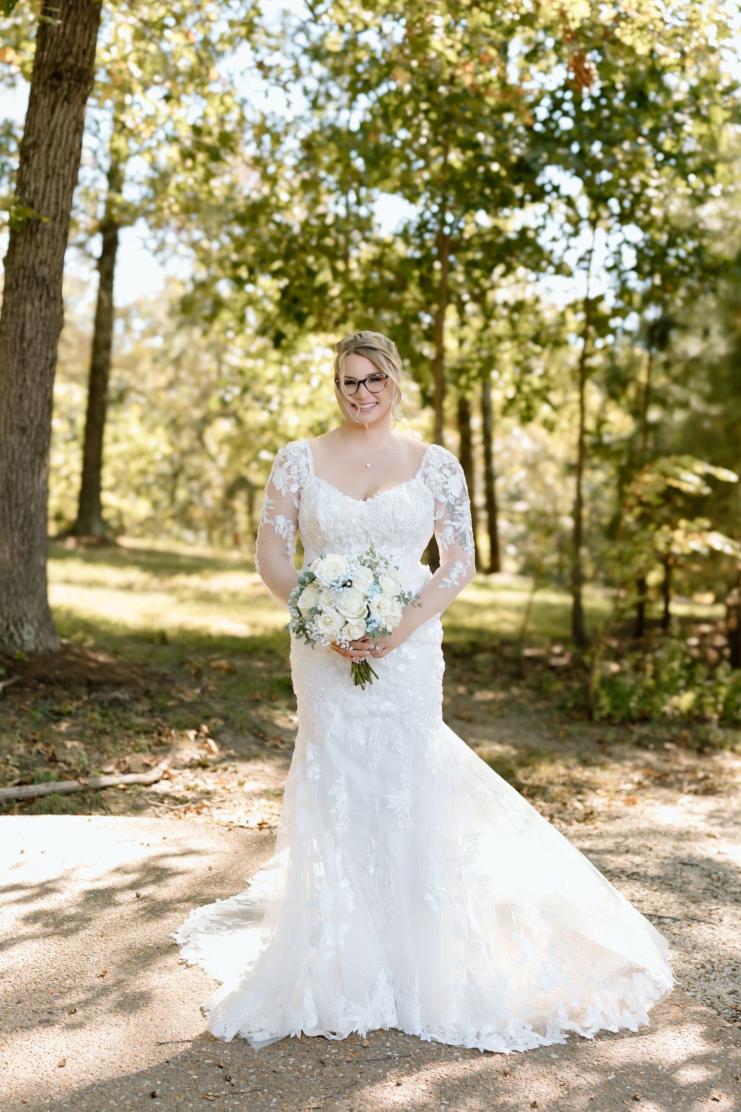 A bride in a flowing white lace gown poses in a sunlit wooded area holding a bouquet of flowers.
