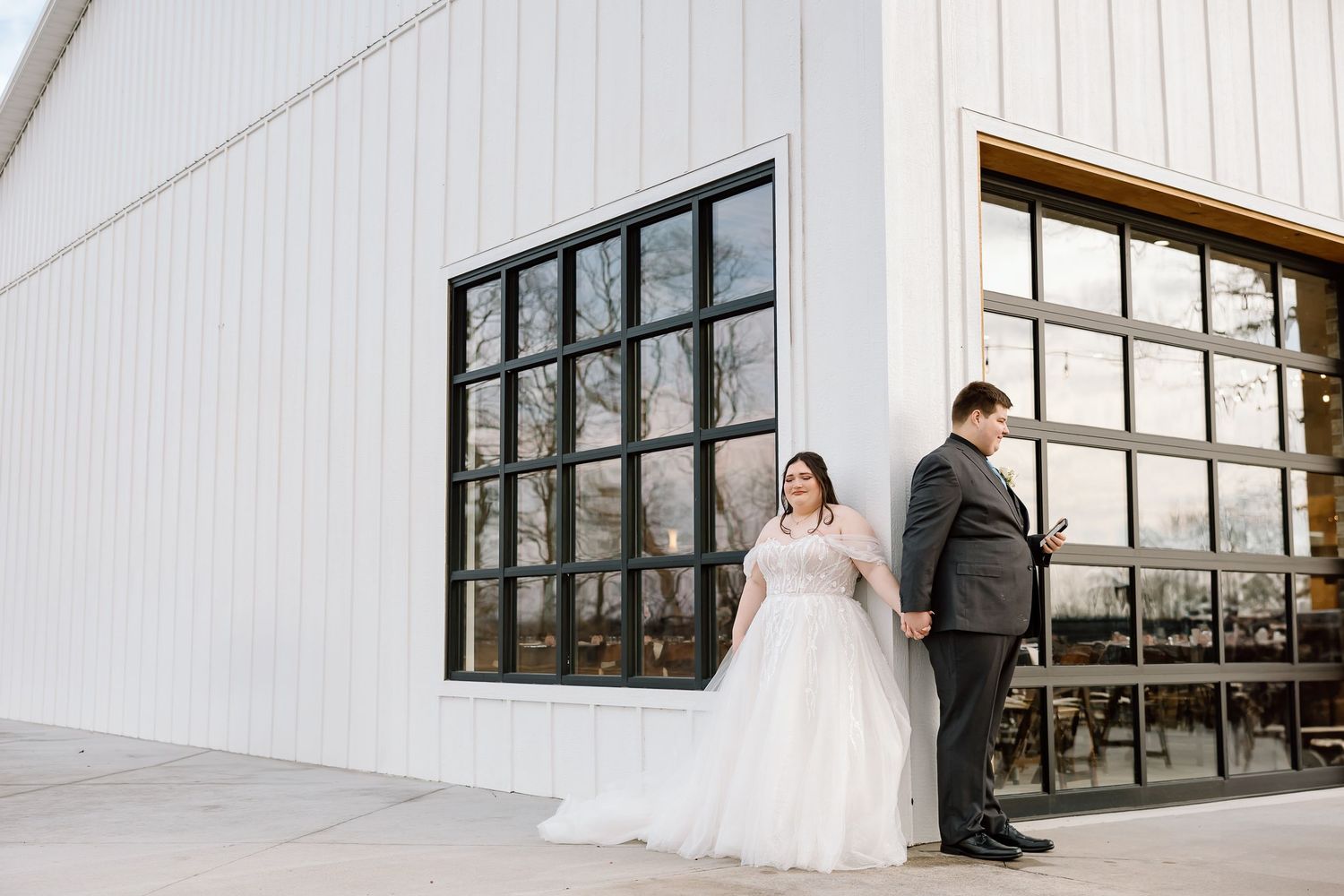 A romantic couple in formal wedding attire stands together against a modern white building exterior.