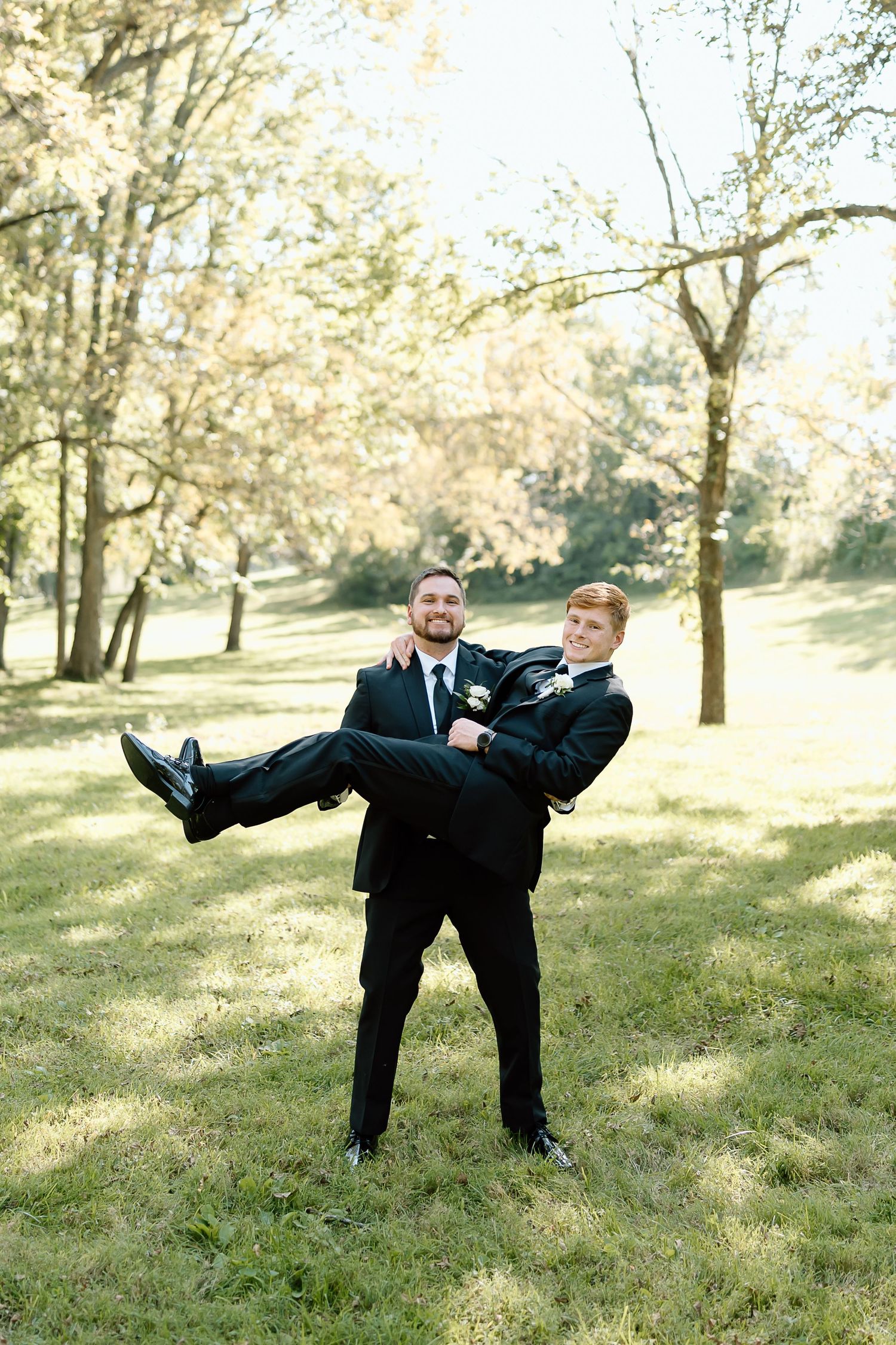 Two people in black suits share a playful moment outdoors as one carries the other in their arms on a sunny day.