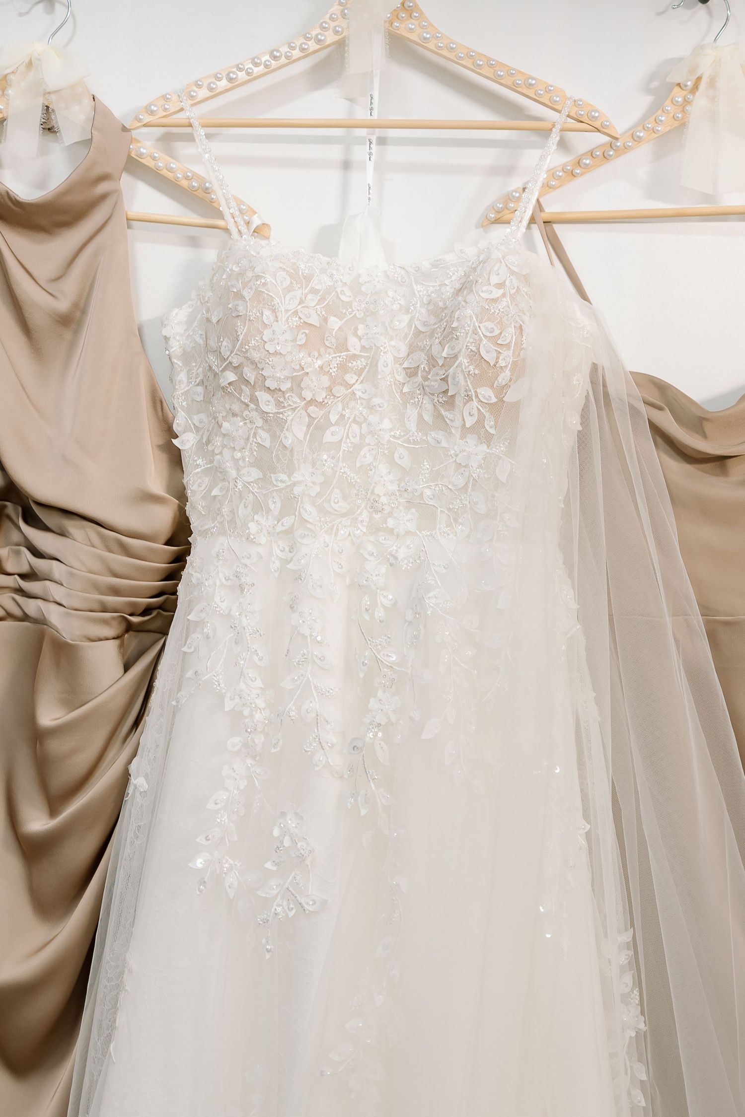 Elegant white wedding dress with delicate lace appliques and floral details hanging on display against beige backdrop.