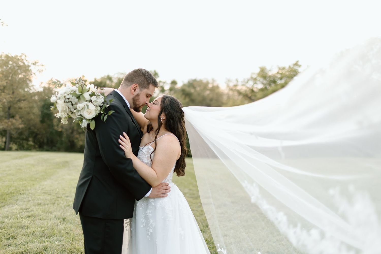 A romantic couple in wedding attire embraces outdoors as the bride's veil flows dramatically in the wind.