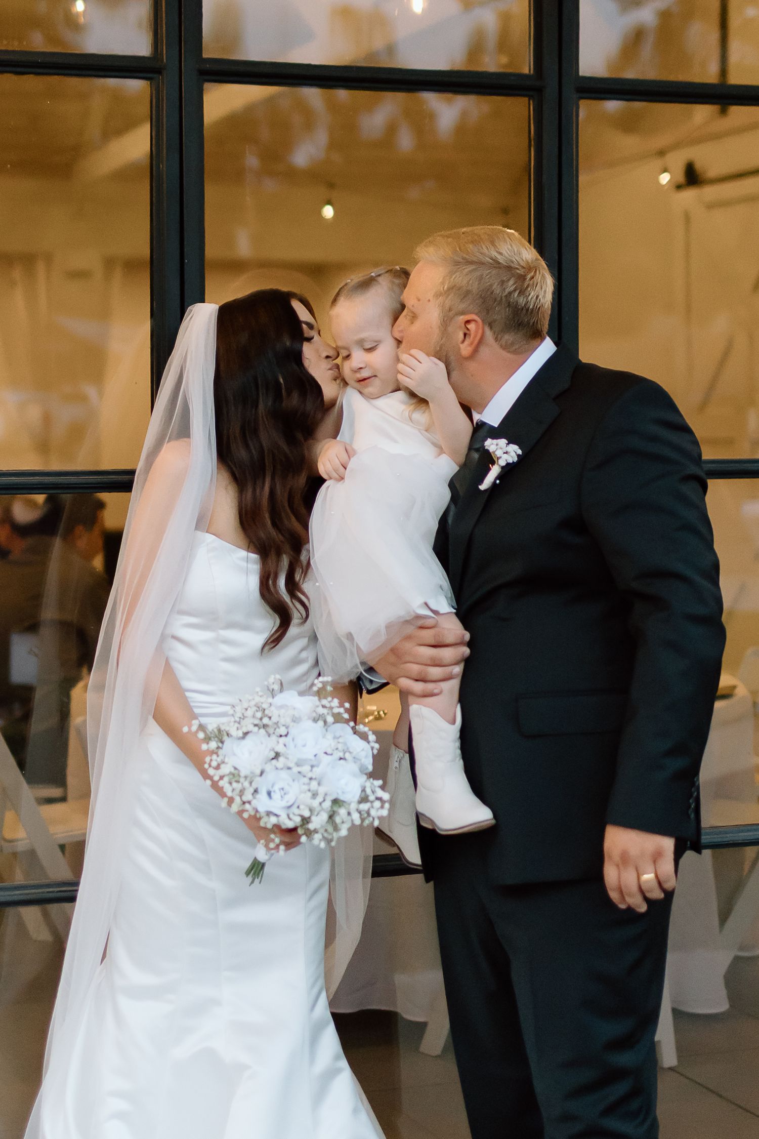 A tender wedding moment captured with a bride in white dress and groom in black suit sharing an embrace with a small child.