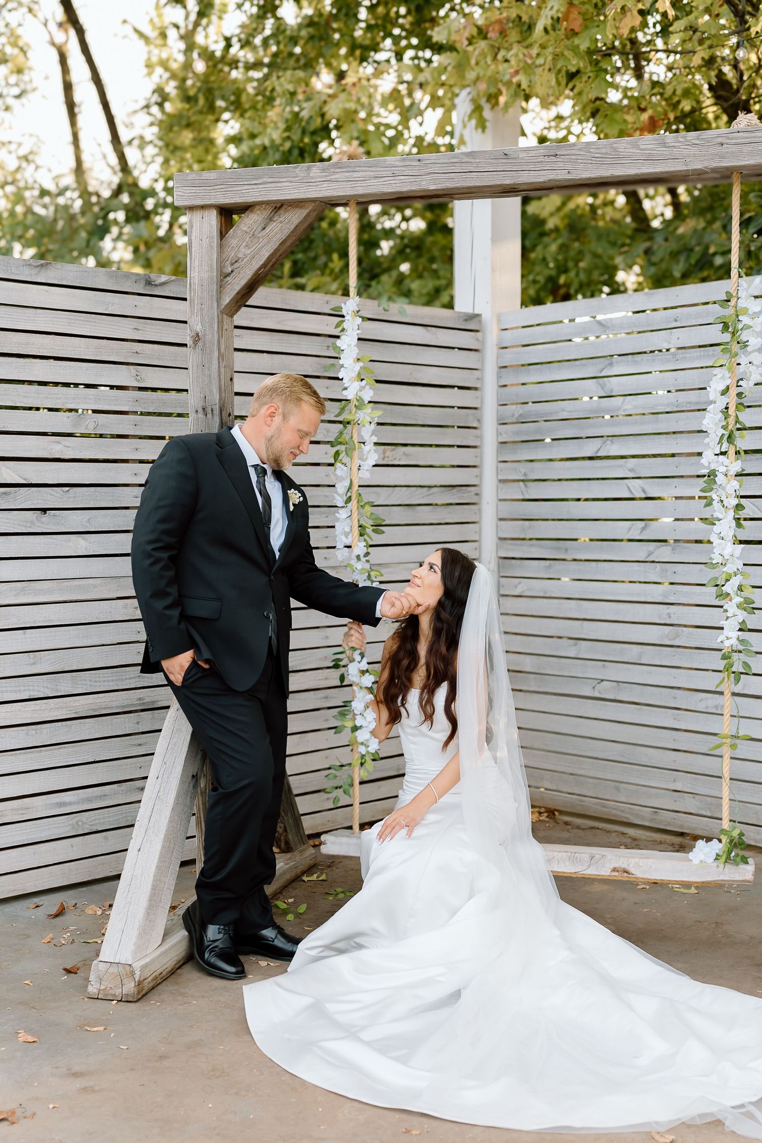 Newlyweds share a romantic moment under a wooden pergola decorated with floral vines and white draping.