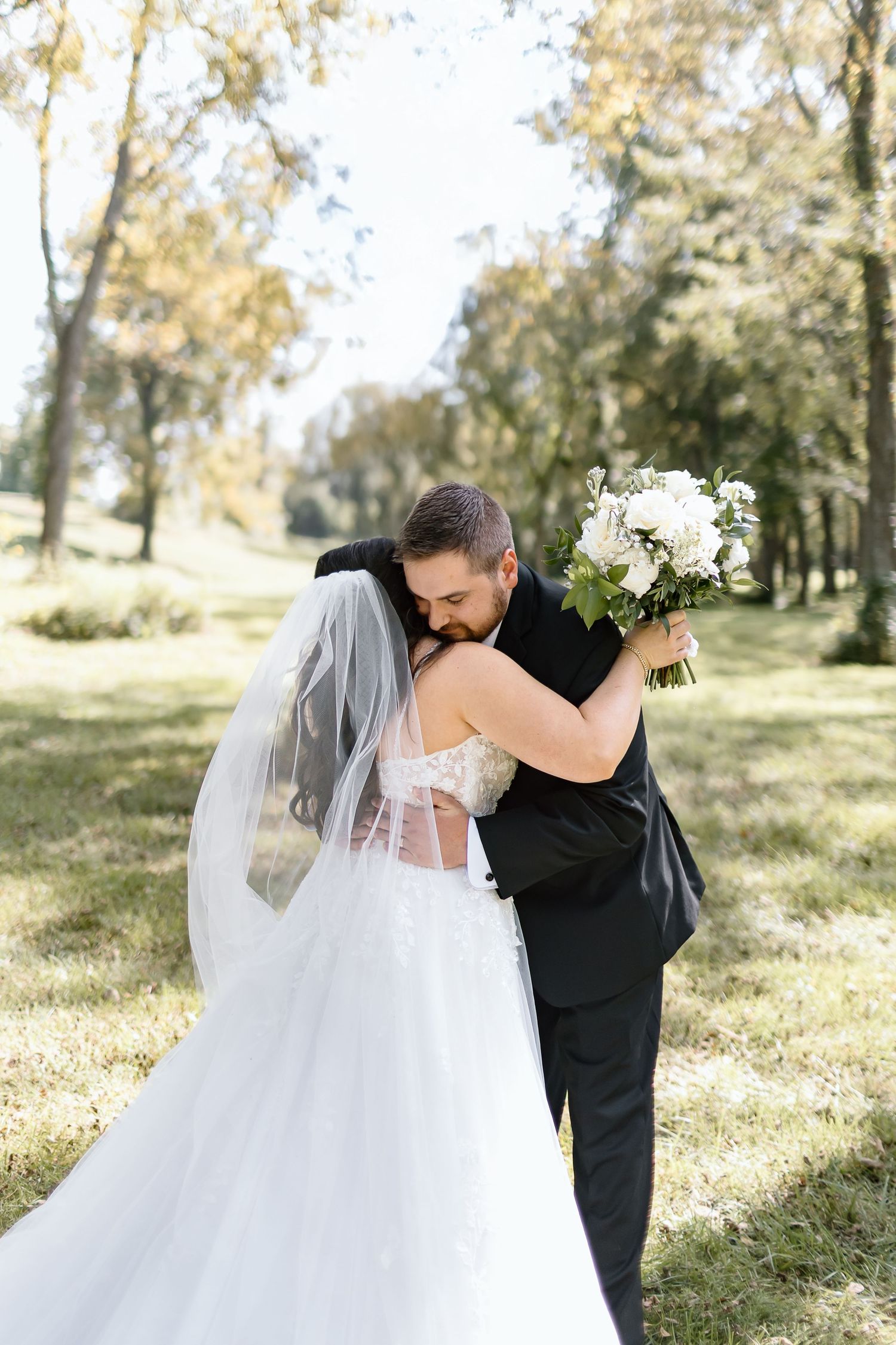 A couple embraces during their outdoor wedding ceremony, with the bride holding a white bouquet in a sunlit wooded setting.