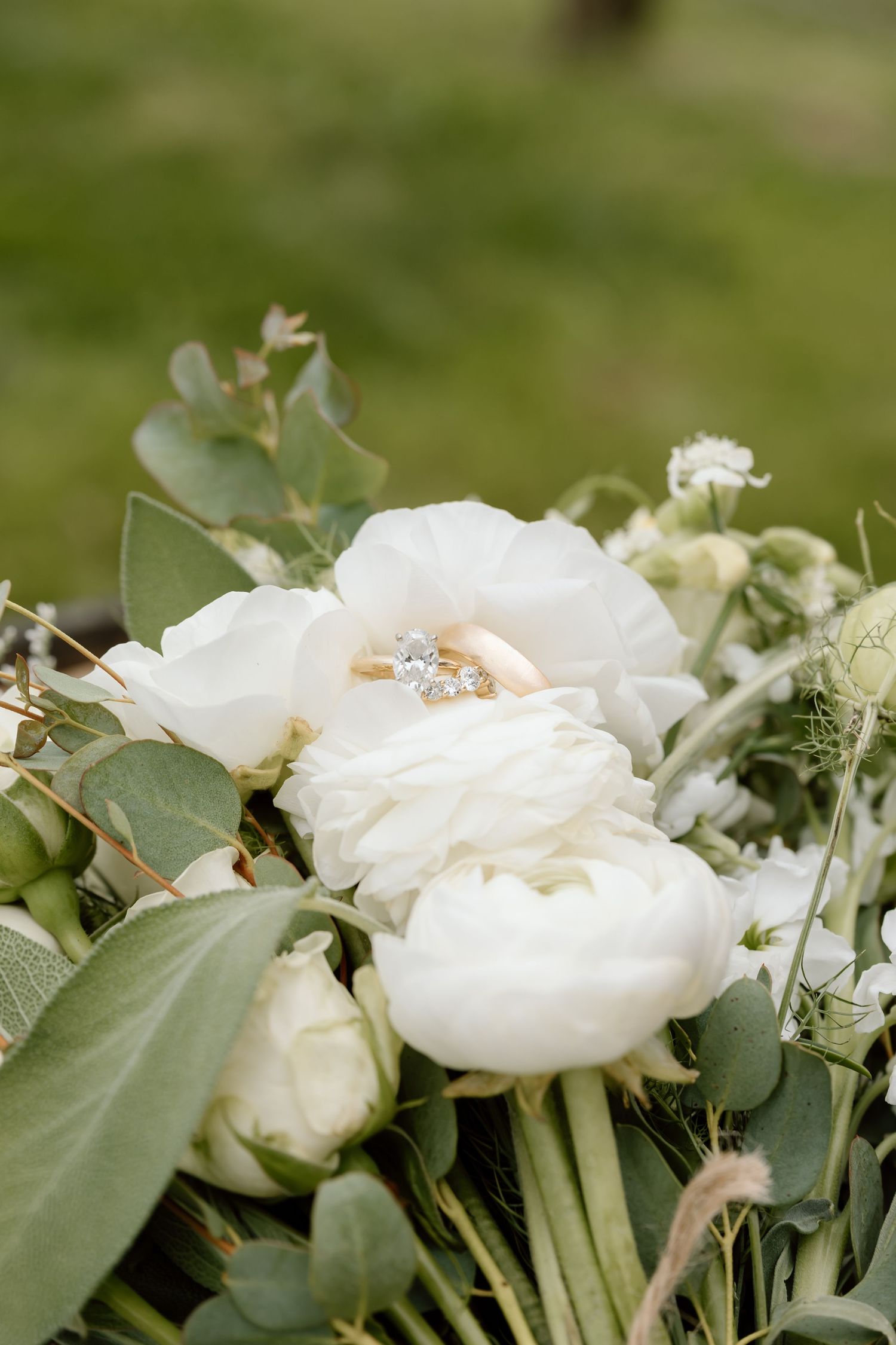 White ranunculus wedding bouquet with gold and diamond rings resting on petals, surrounded by eucalyptus greenery.