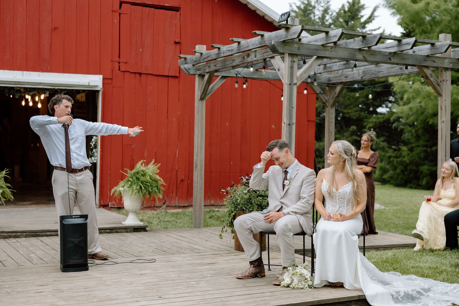 Outdoor barn wedding ceremony with officiant gesturing to couple seated under wooden pergola near red barn.