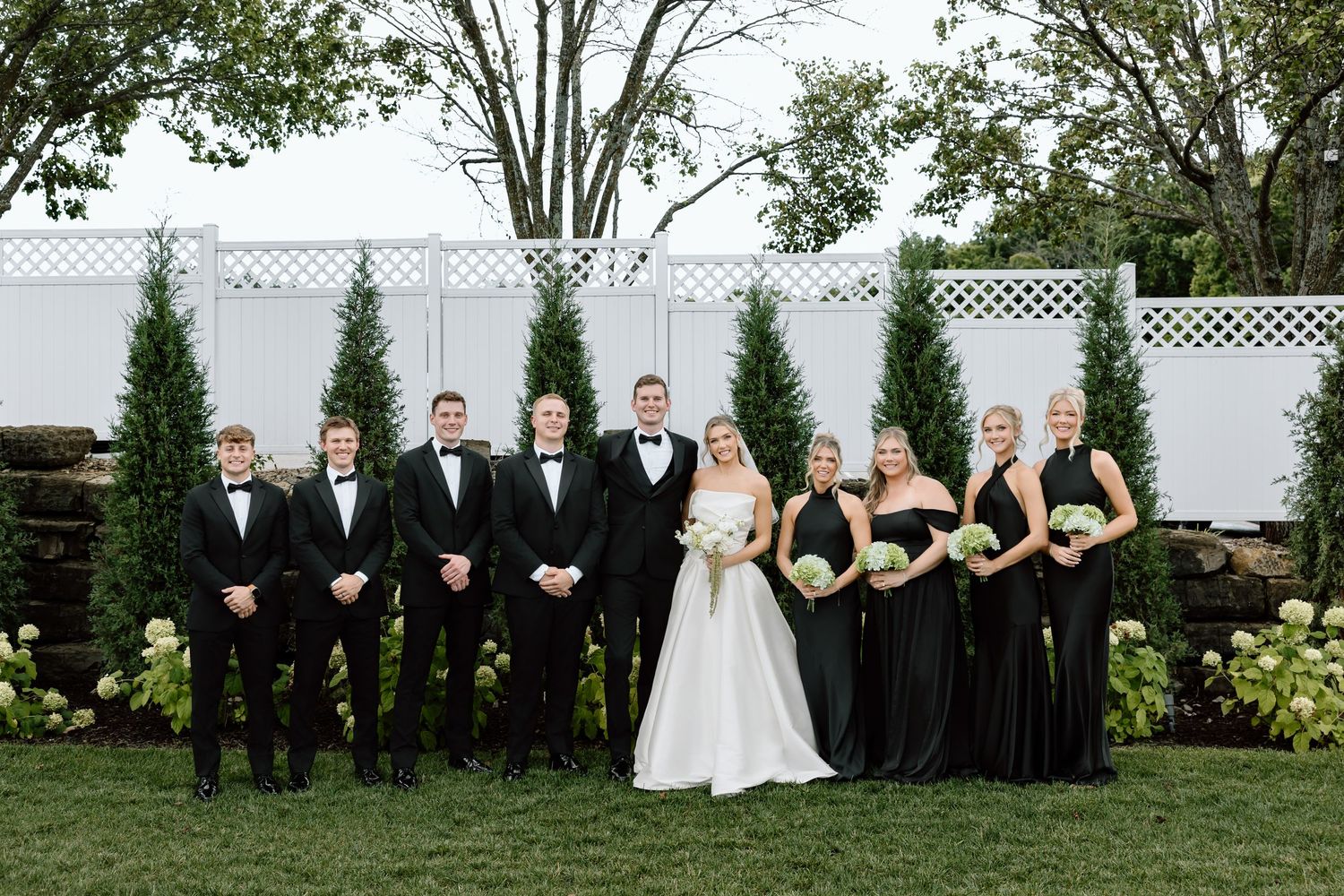 Wedding party in black attire posing outdoors with bride and groom in front of evergreen trees.
