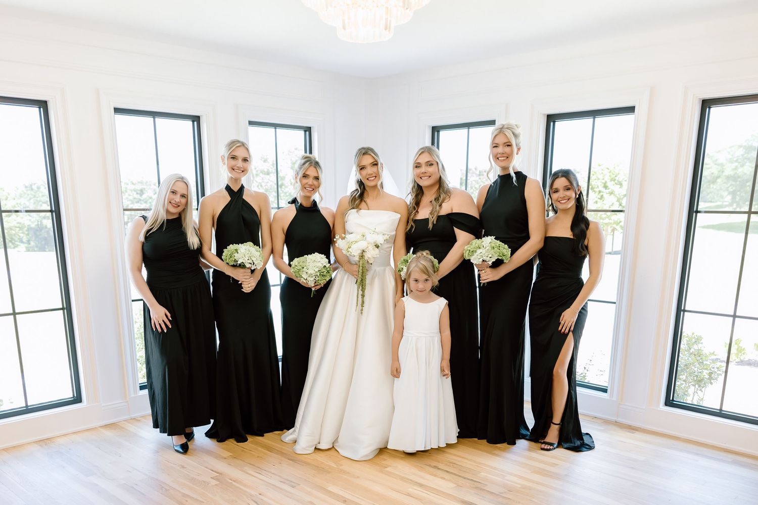 Bride in white gown with bridesmaids in black dresses and flower girl posing indoors with bouquets.