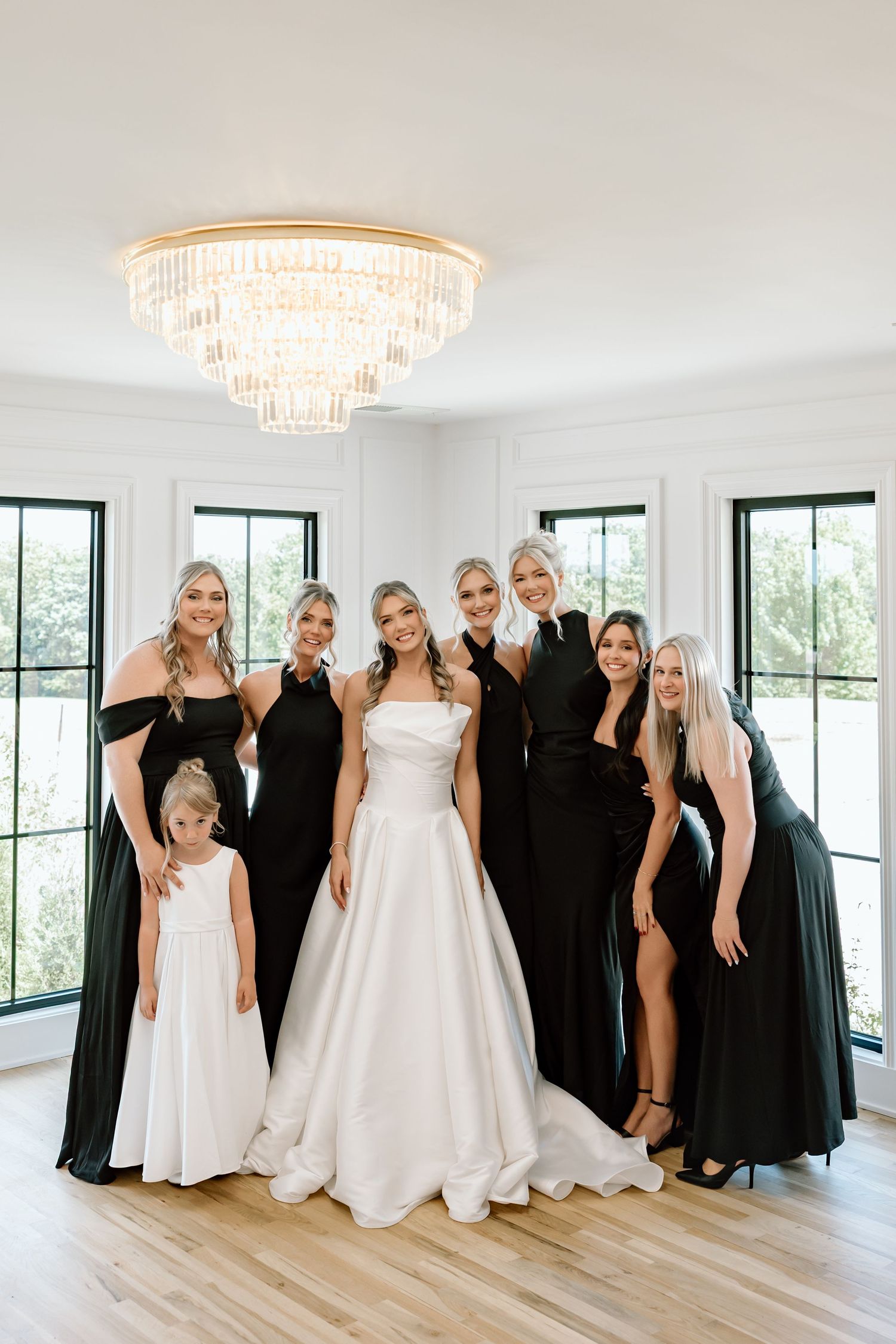 Bride in white gown poses with bridesmaids in black dresses and flower girl under elegant chandelier.