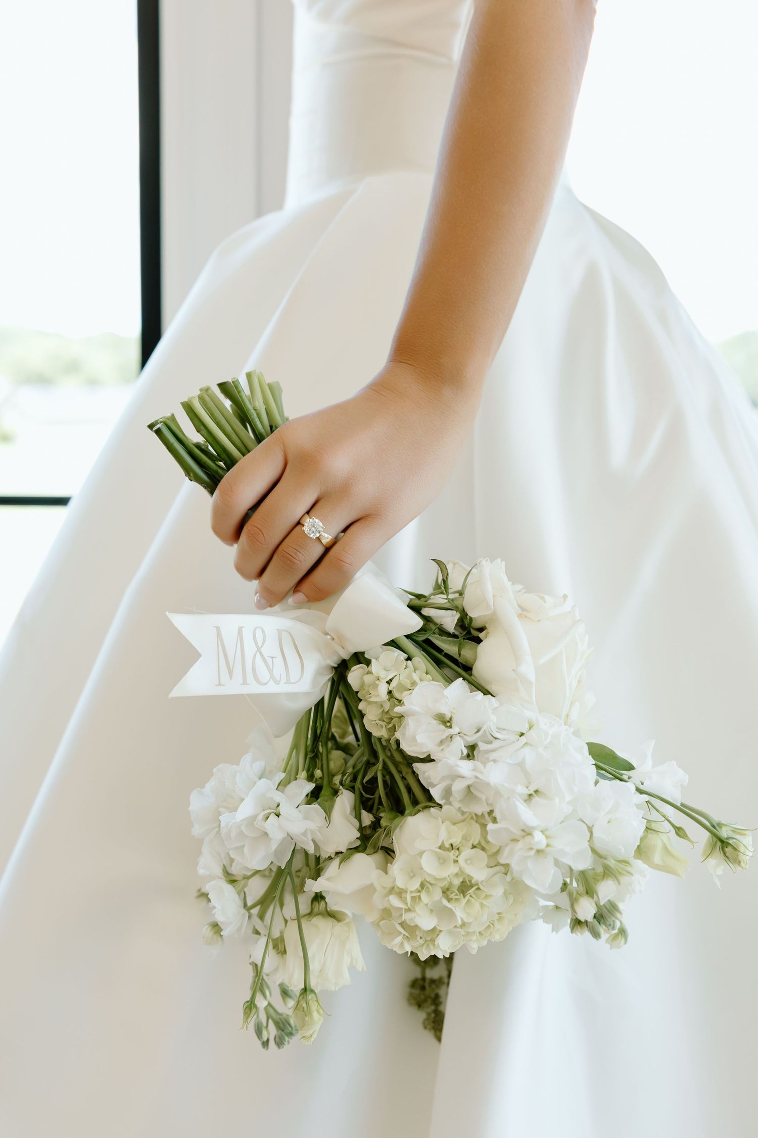 Bride holding white floral bouquet with M&Y ribbon charm, wearing diamond engagement ring in elegant satin wedding gown.