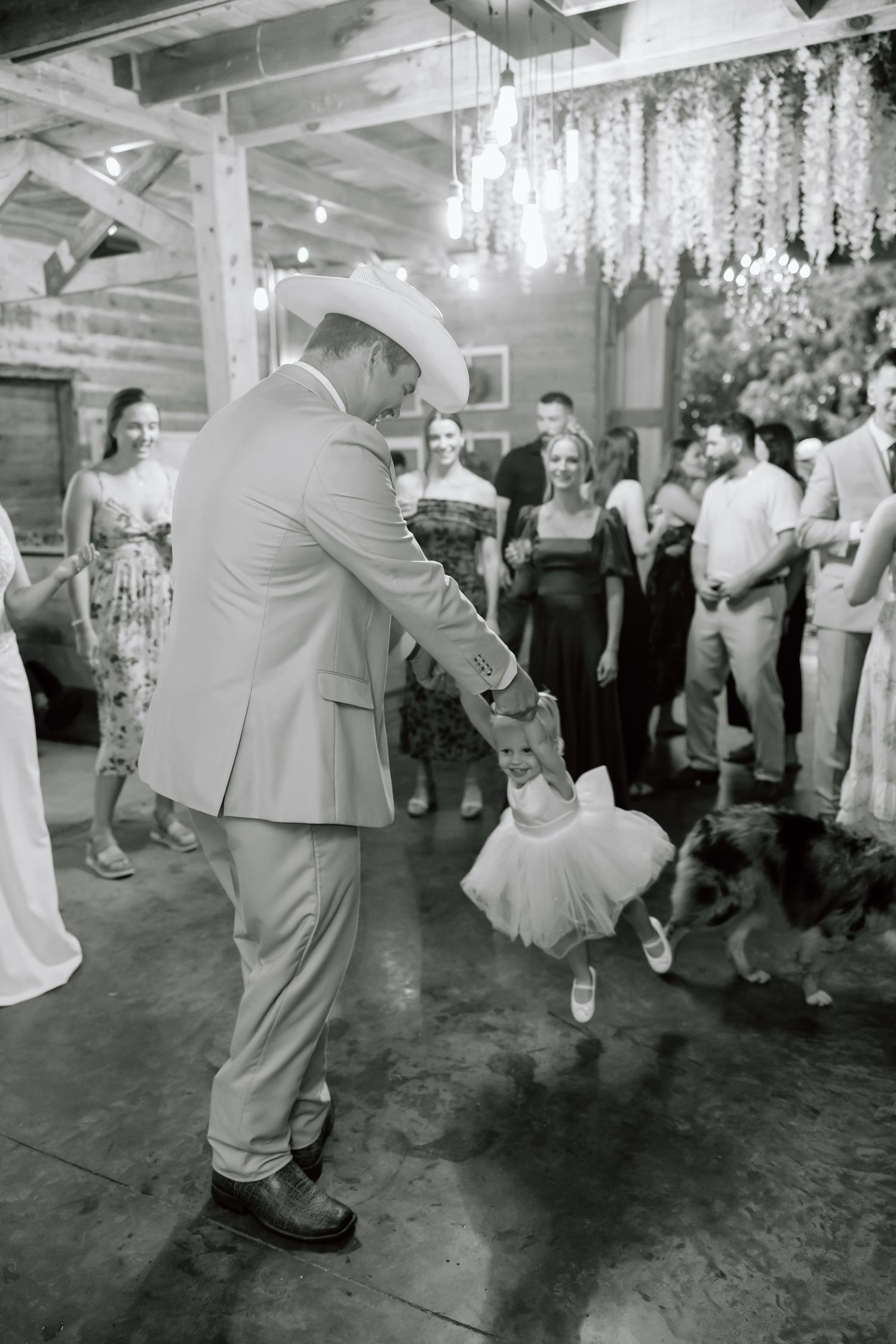 Groom in cowboy hat twirls young flower girl in tutu on dance floor at rustic barn wedding reception.