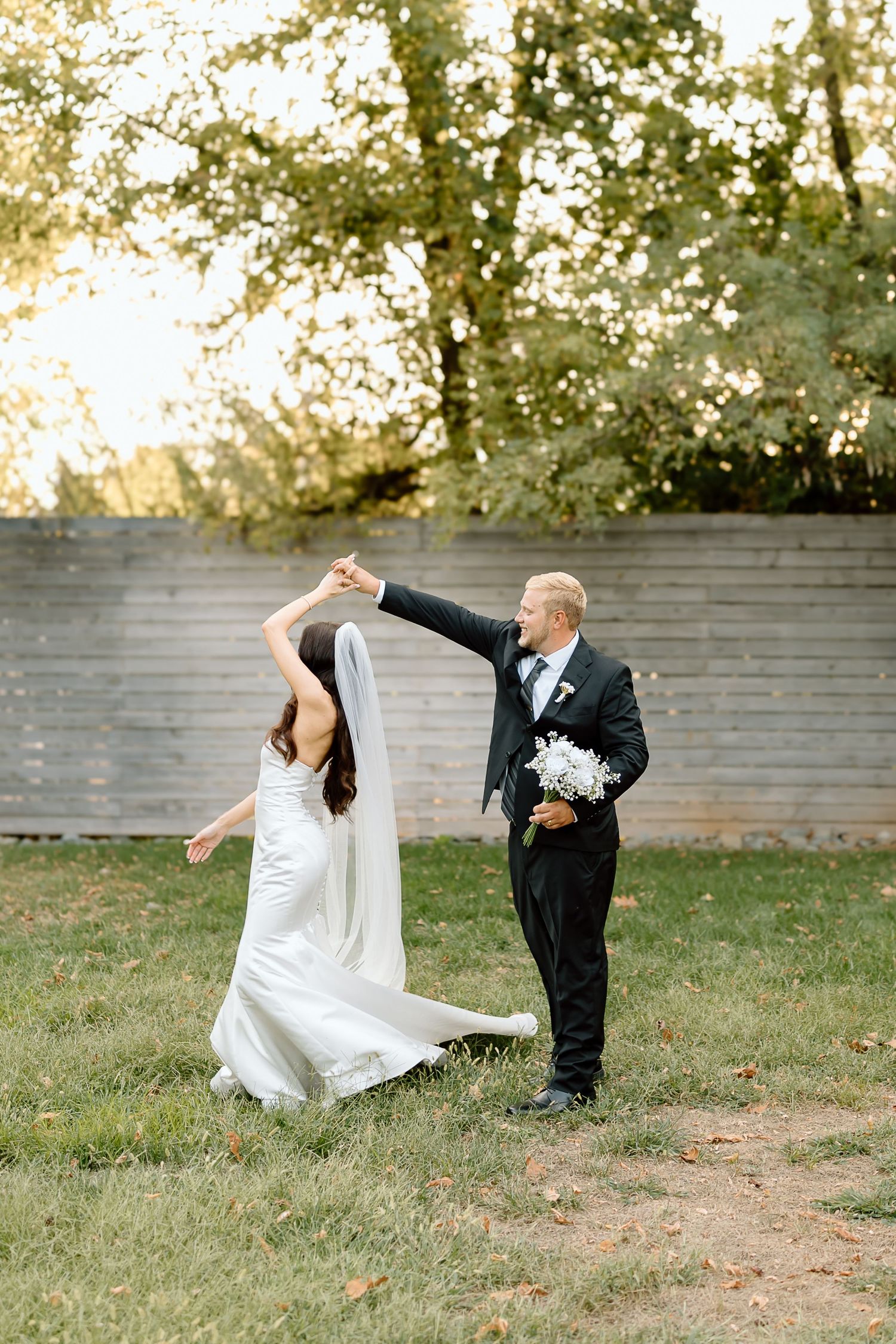 A couple in wedding attire playfully dances and twirls together on a grassy lawn with a stone wall backdrop.