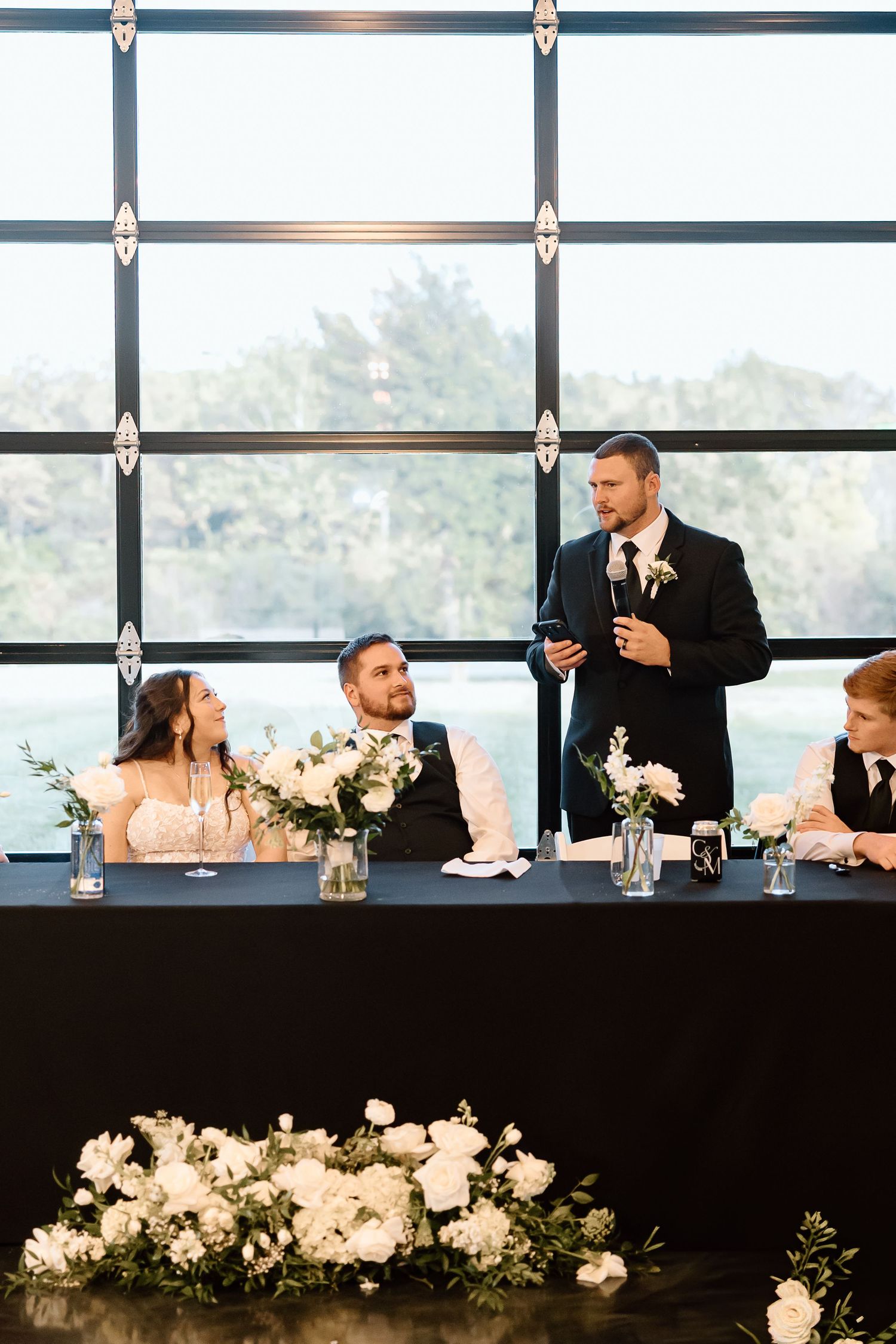 Wedding party seated at head table during reception with decorative white floral arrangements in foreground.