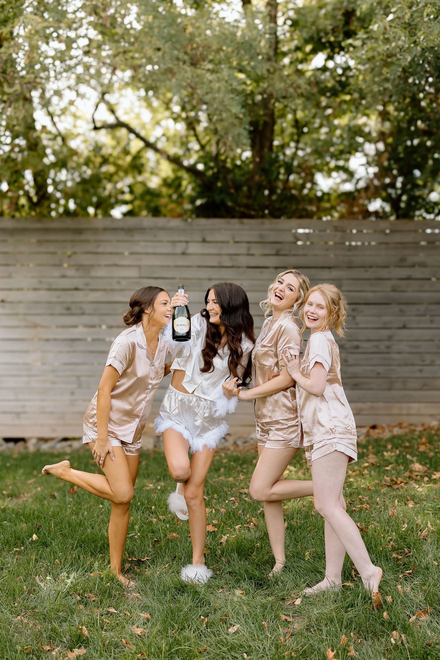 A group of friends in light colored outfits play together on grass near a wooden fence in a backyard setting.