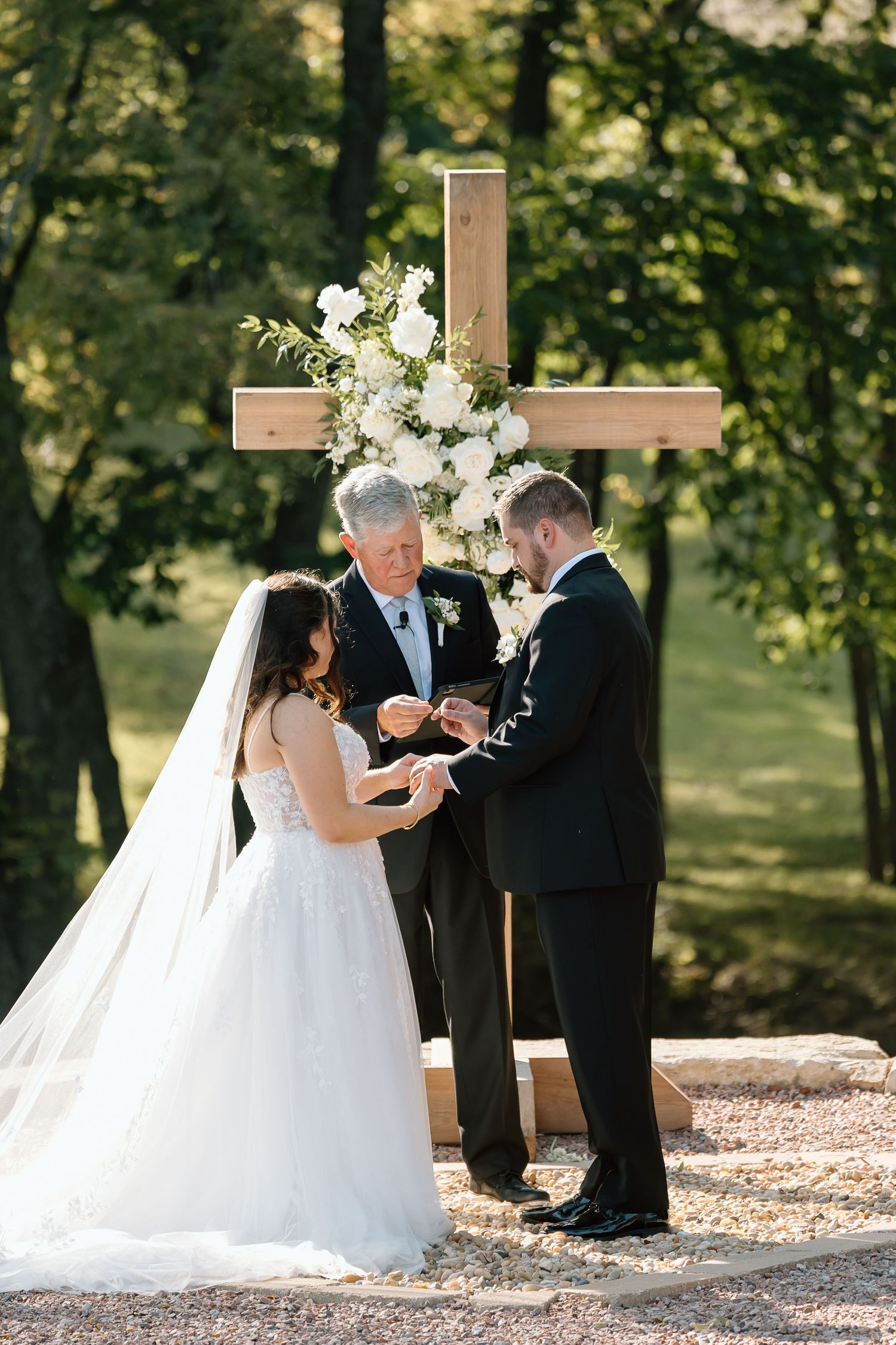 Couple exchanges wedding vows during outdoor ceremony with wooden cross and lush greenery in background.